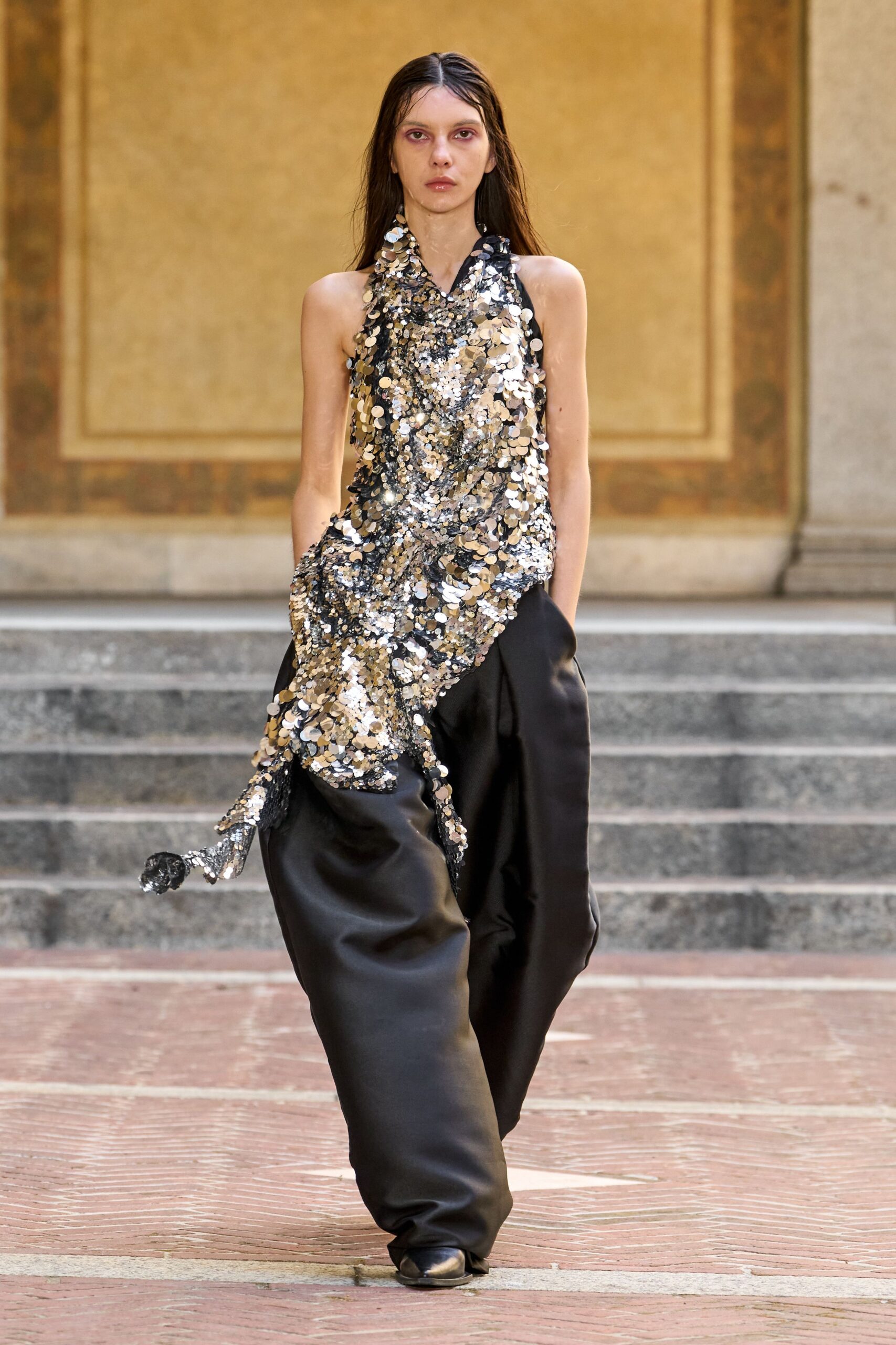 Halter-draped black mesh buried under oversized silver paillettes. Black satin trousers cut full and puddling over the shoe. Square-toe black flats, just peeking from the hem. Crossing a museum courtyard after the gala has spilled outside.