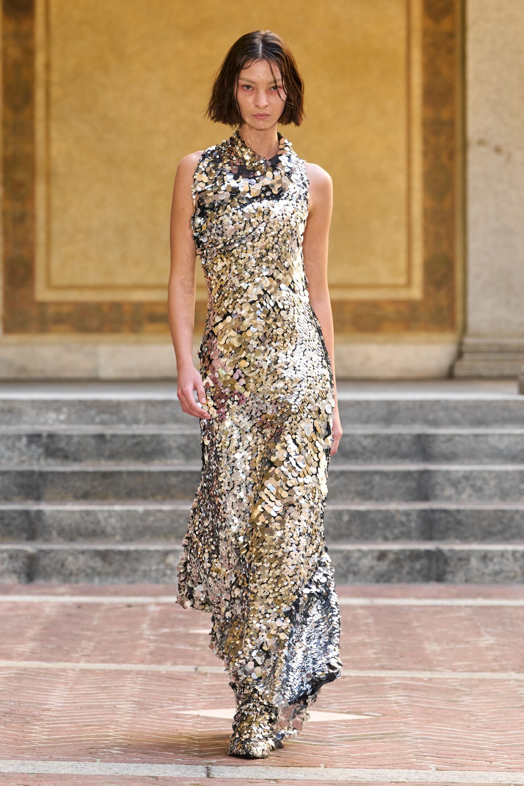 Floor-length silver paillette gown, sleeveless and densely scaled from collar to hem. Matching sequin boots nearly swallowed by the dragging hem. Crossing a palazzo courtyard after midnight, still throwing back every flash.
