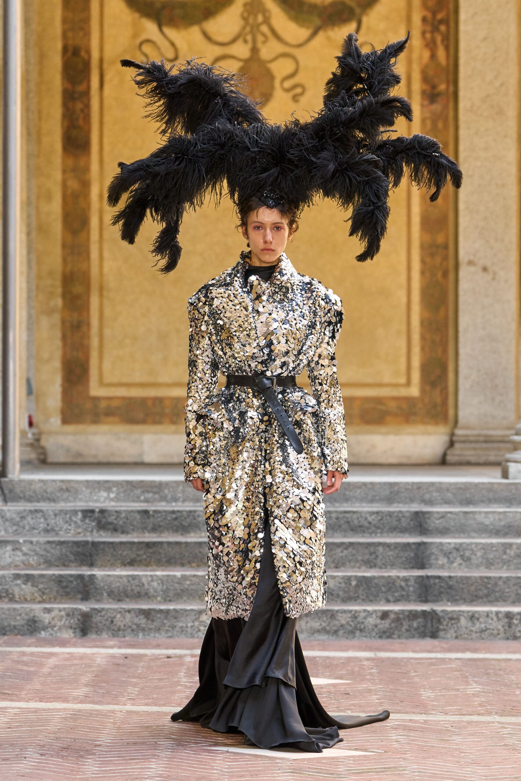 Black ostrich plumes branching into huge antler-like sprays. Long silver paillette coat, broad lapels and one shoulder jutting higher. Black leather belt with the tongue hanging past the buckle. Black satin skirt pooling into a fishtail train. Crossing a frescoed courtyard just before the masked ball begins.