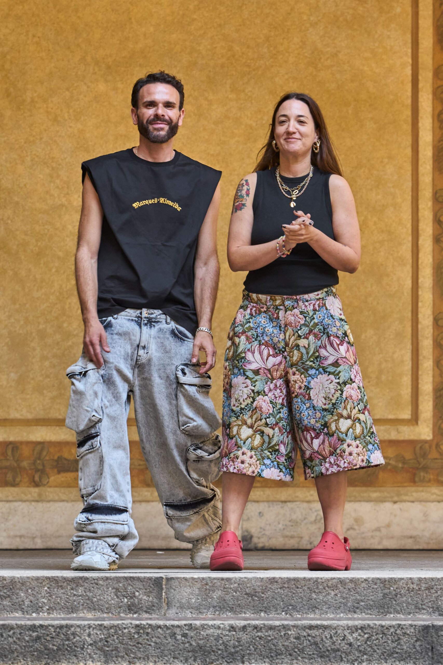 Black cotton muscle tee with a stiff, apronlike bib front. Acid-wash denim cargo jeans with bulging pouch pockets at the thighs. Off-white sneakers swallowed by the pooled hems. Walking down municipal stone steps after clapping through the student showcase.