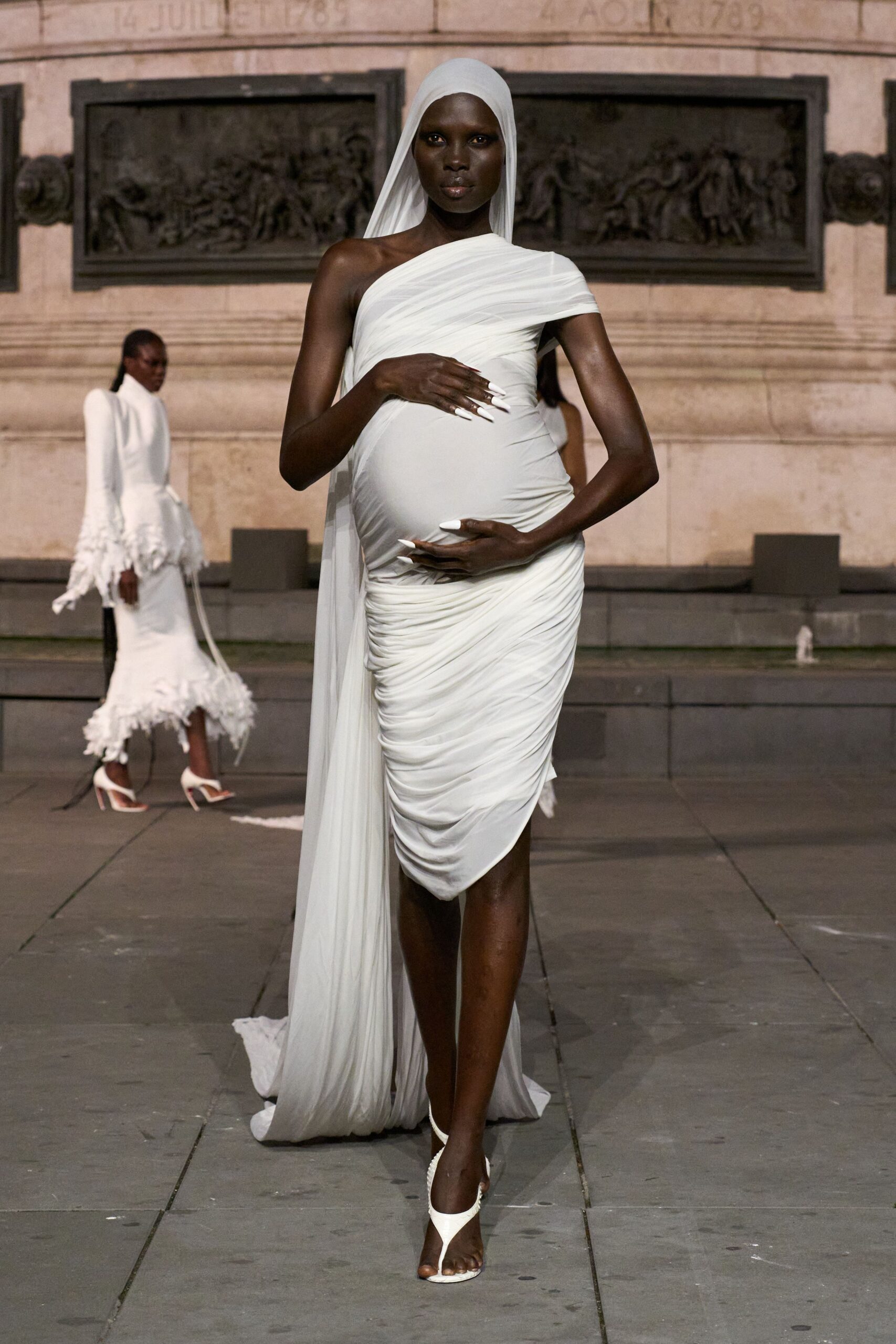 Long white jersey veil falling in a narrow column to the ground. Ivory stretch dress, densely ruched across one shoulder and over the belly. White thong heels with a slim curved stiletto. Long white pointed acrylics. Crossing a stone square after the civil ceremony, dusk still holding heat.