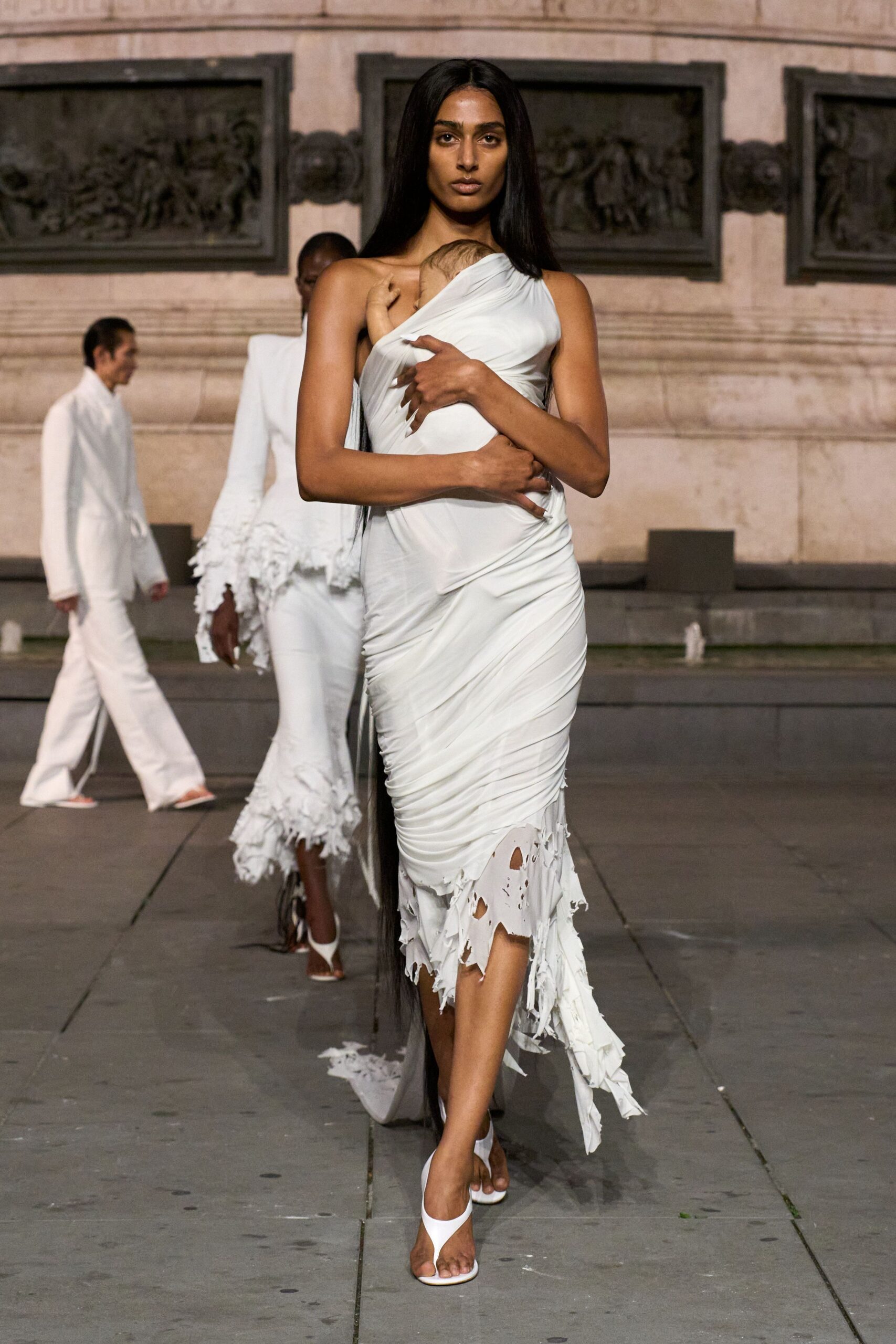 White jersey dress wrapped one-shoulder around a swaddled baby, ruched to the hip. Shredded gauze godet trailing behind, punched through with ragged holes. White thong heels with a broad curved strap. Crossing a stone square at dawn after the christening ran all night.