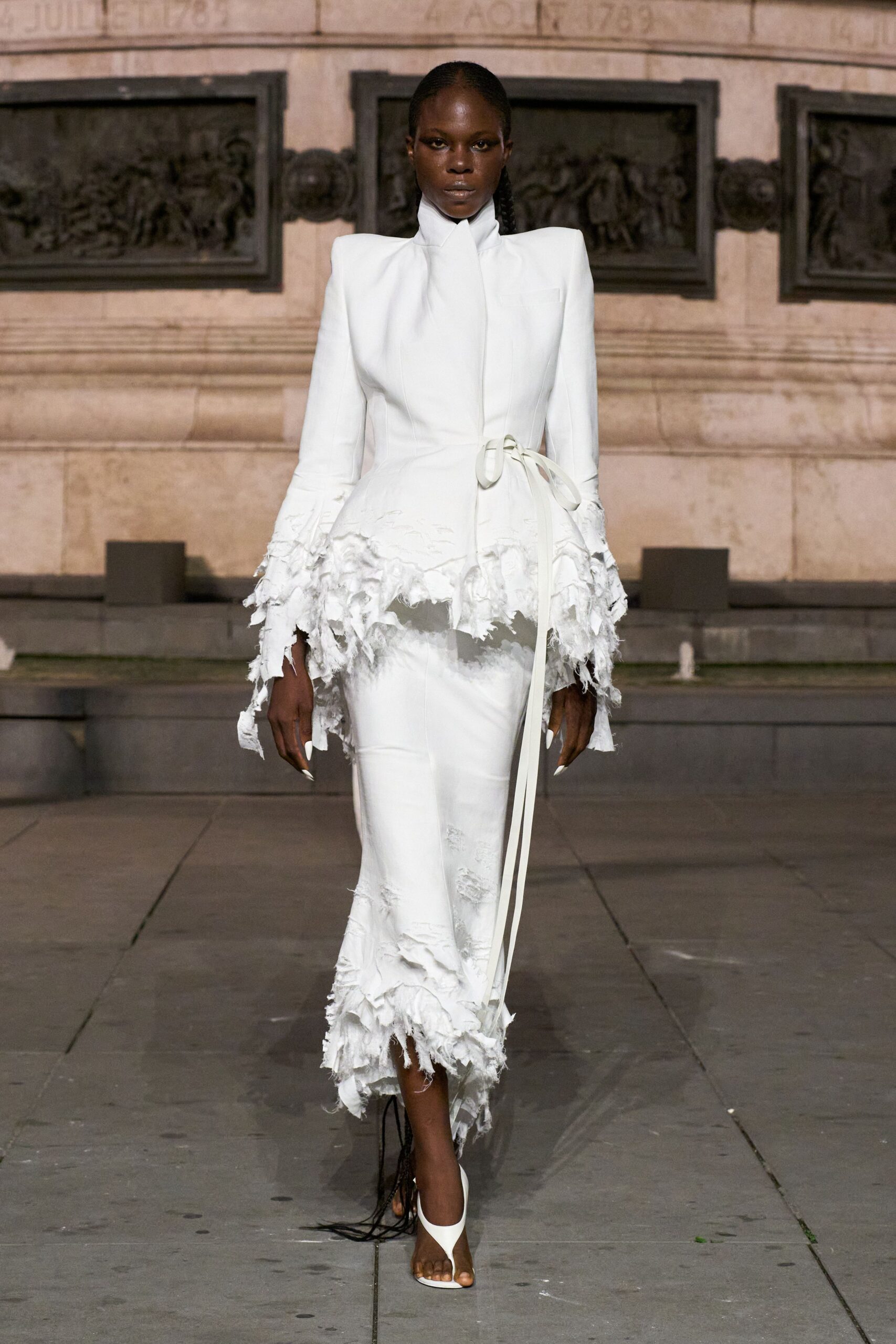 Ivory fitted jacket with a stand collar and shredded peplum hem. Narrow matching skirt, ripped away in cloudy bands above the ankle. White thong heels with black fringe trailing off the back. Crossing a stone square just after the wedding crowd has gone.