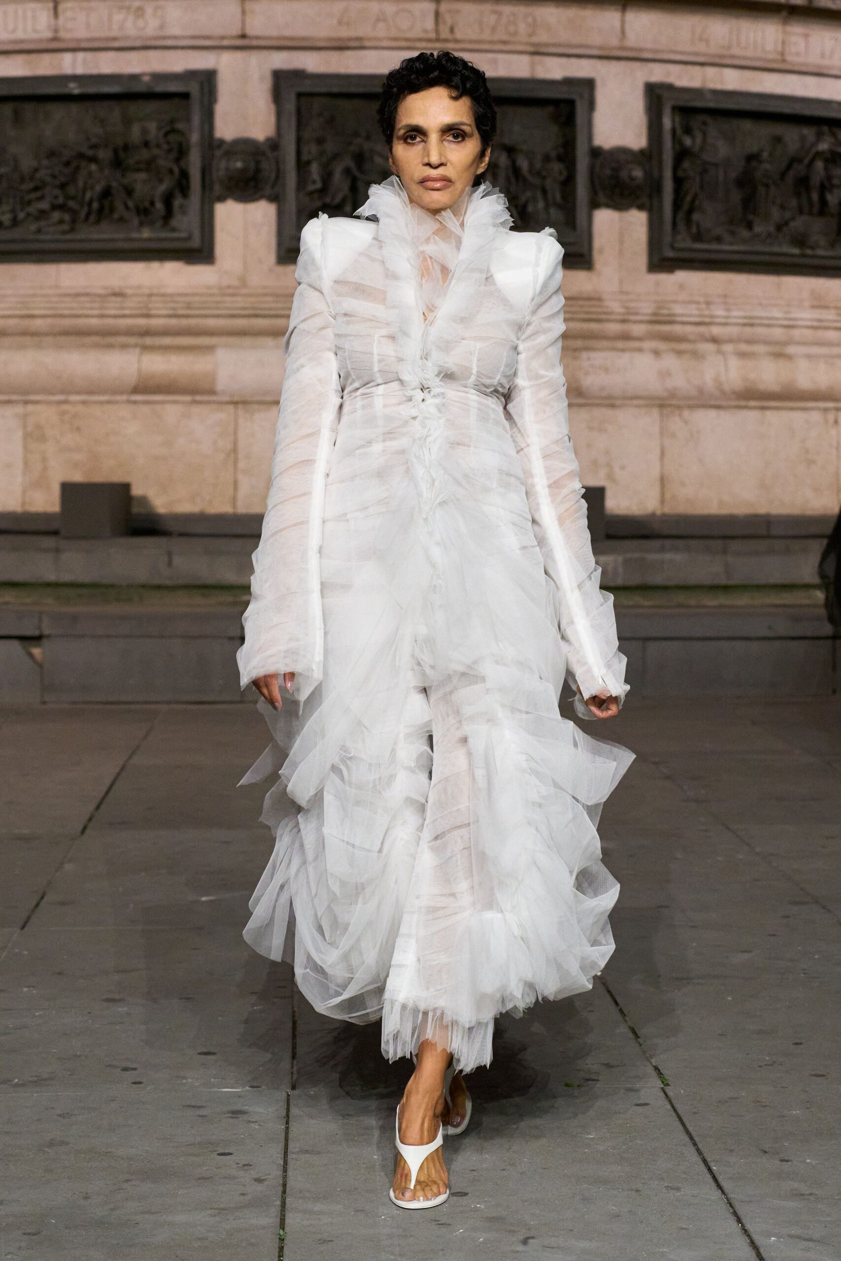 Sheer white tulle gown ruched in spirals, ruff climbing the throat. White thong mules with a clear lucite heel. Crossing a stone courtyard after the ceremony, tulle still damp at the hem.