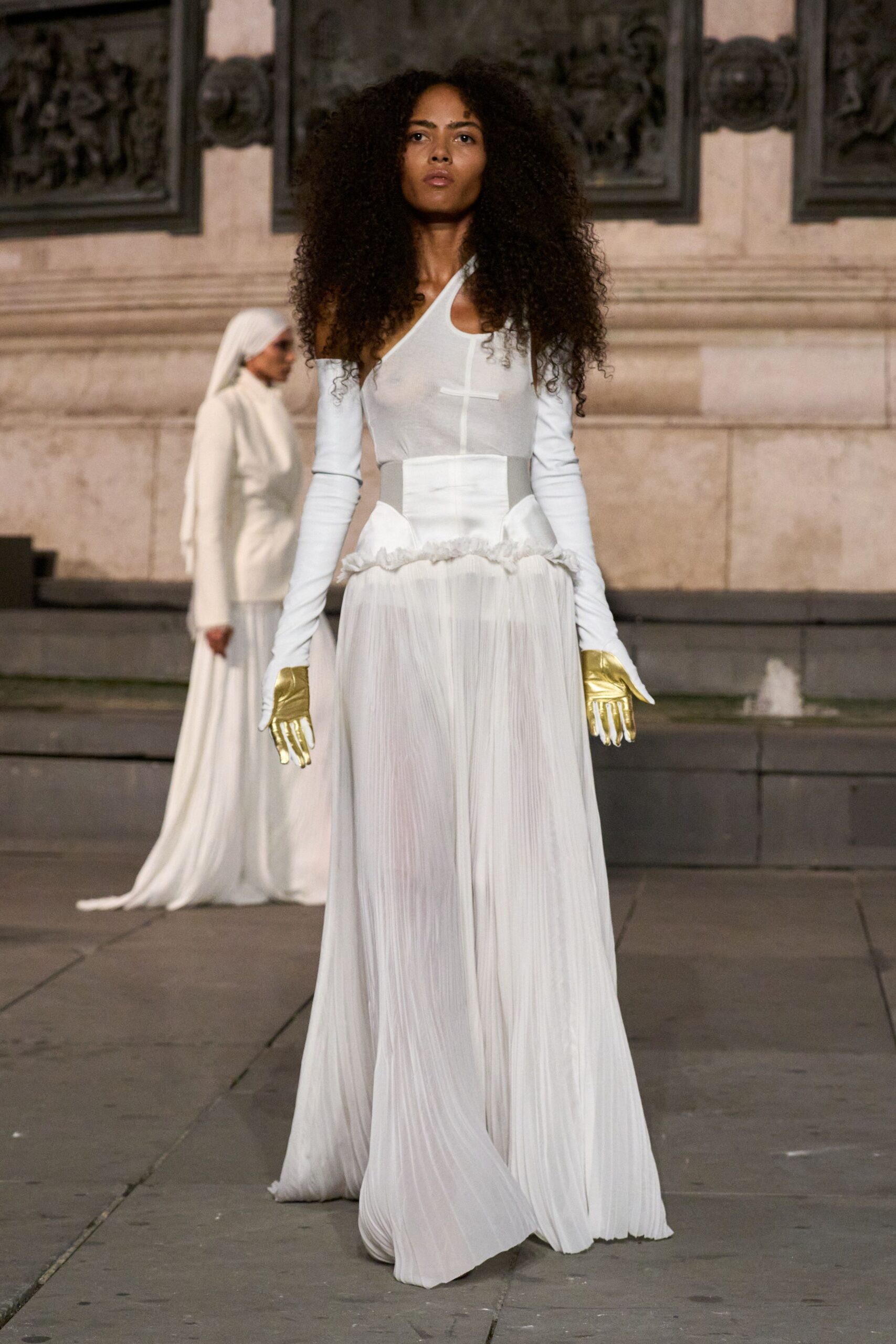 Sheer white one-shoulder bodice with a narrow crossbar seam at the chest. Long white sleeves ending in metallic gold glove hands. Floor-grazing white plisse trousers hidden beneath a peplum of soft ruffles. Crossing a stone square just after the fountain switches on.