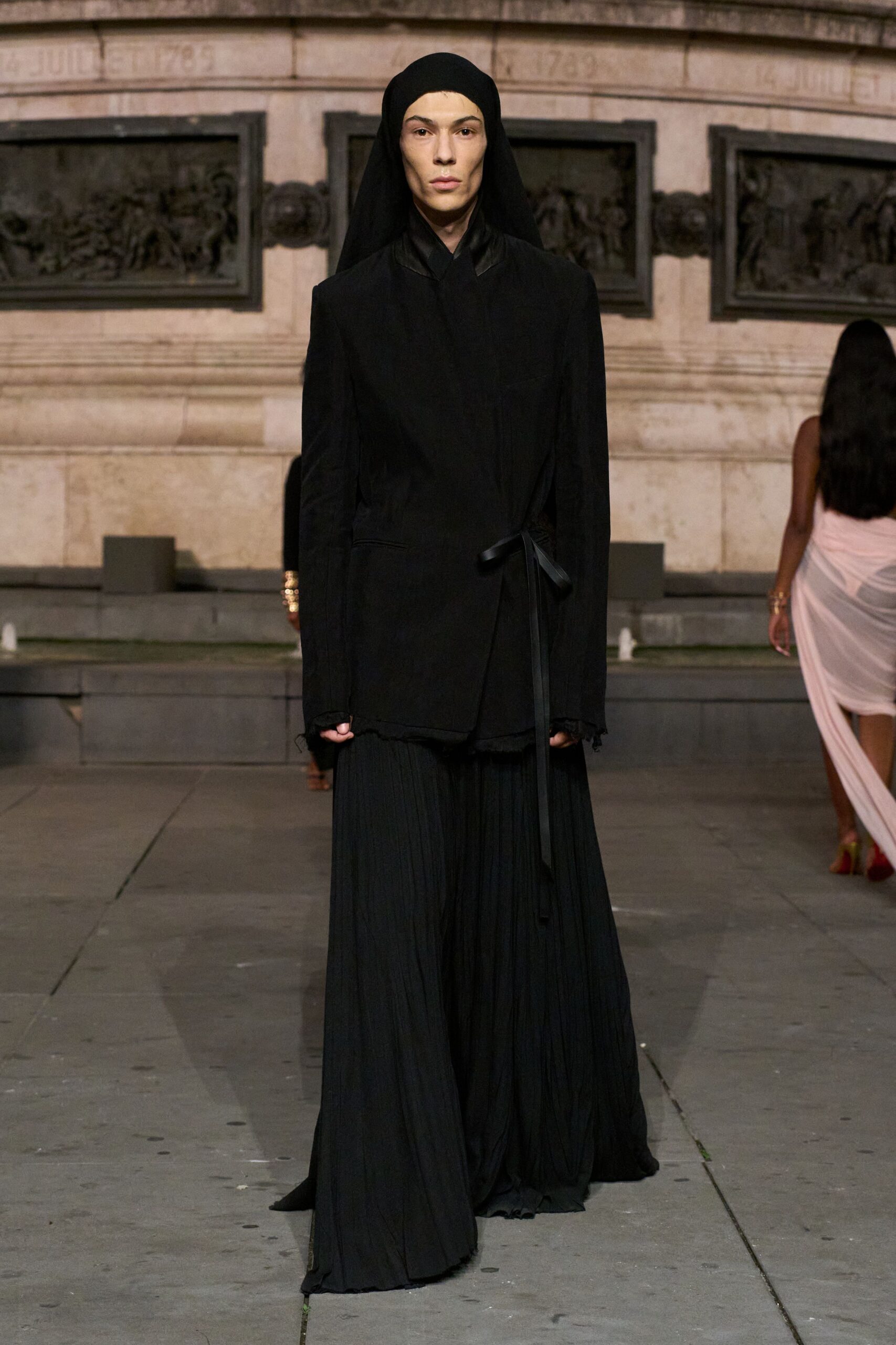 Black jersey headscarf falling in a long veil behind the shoulders. Matte black wrap jacket, satin collar and a narrow side tie. Floor-pooling pleated trousers with a softly crinkled drape. Black toes barely showing. Crossing a stone square just after the fountain shuts off.