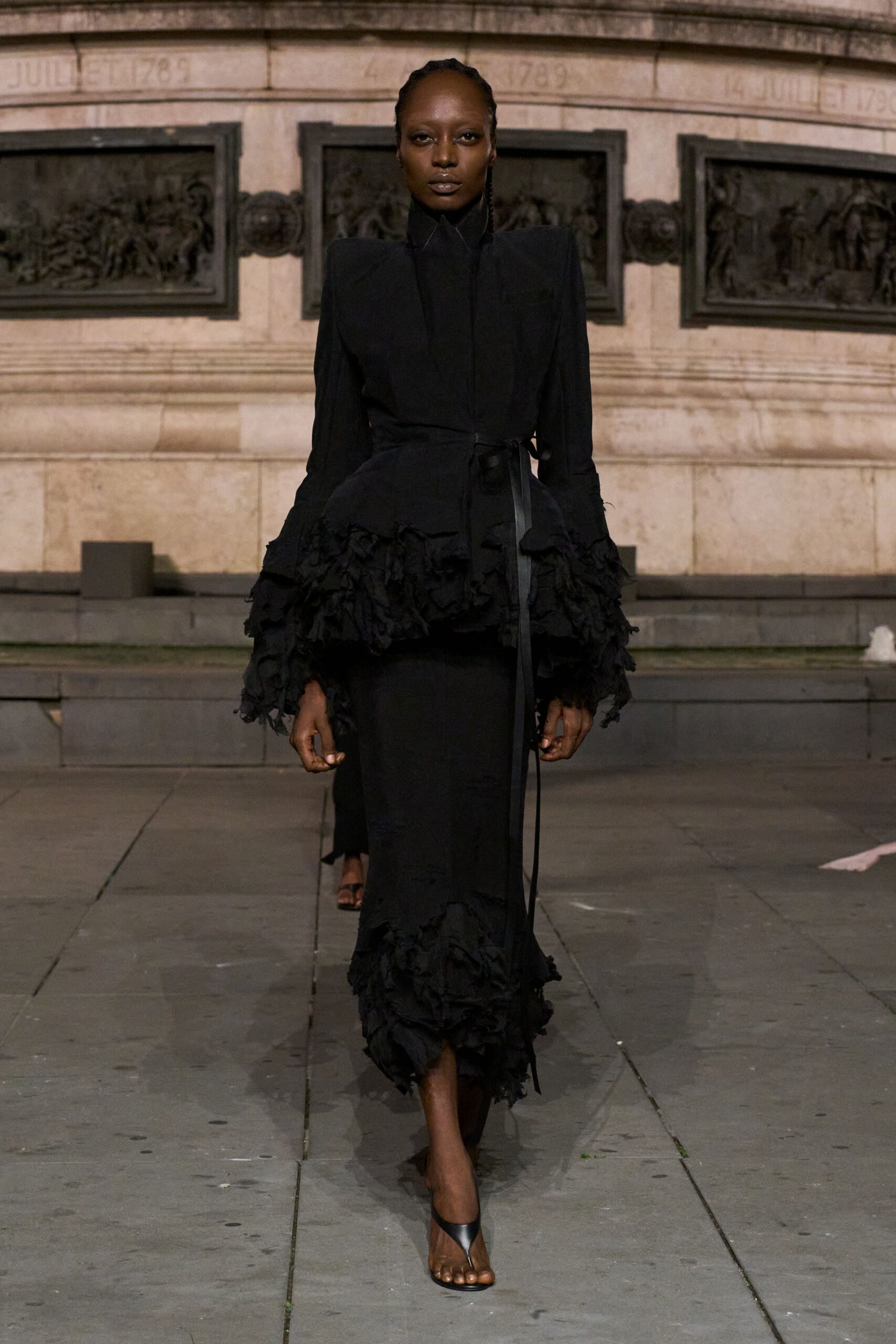 Black coatdress with a blade-sharp stand collar and ribbon-tied waist. Tiered ruffles shredding outward at the sleeves and dropped peplum. Narrow black skirt ending in the same ragged flounced hem. Black thong-strap heels. Crossing a stone square after rain, monument bronze still holding the heat.