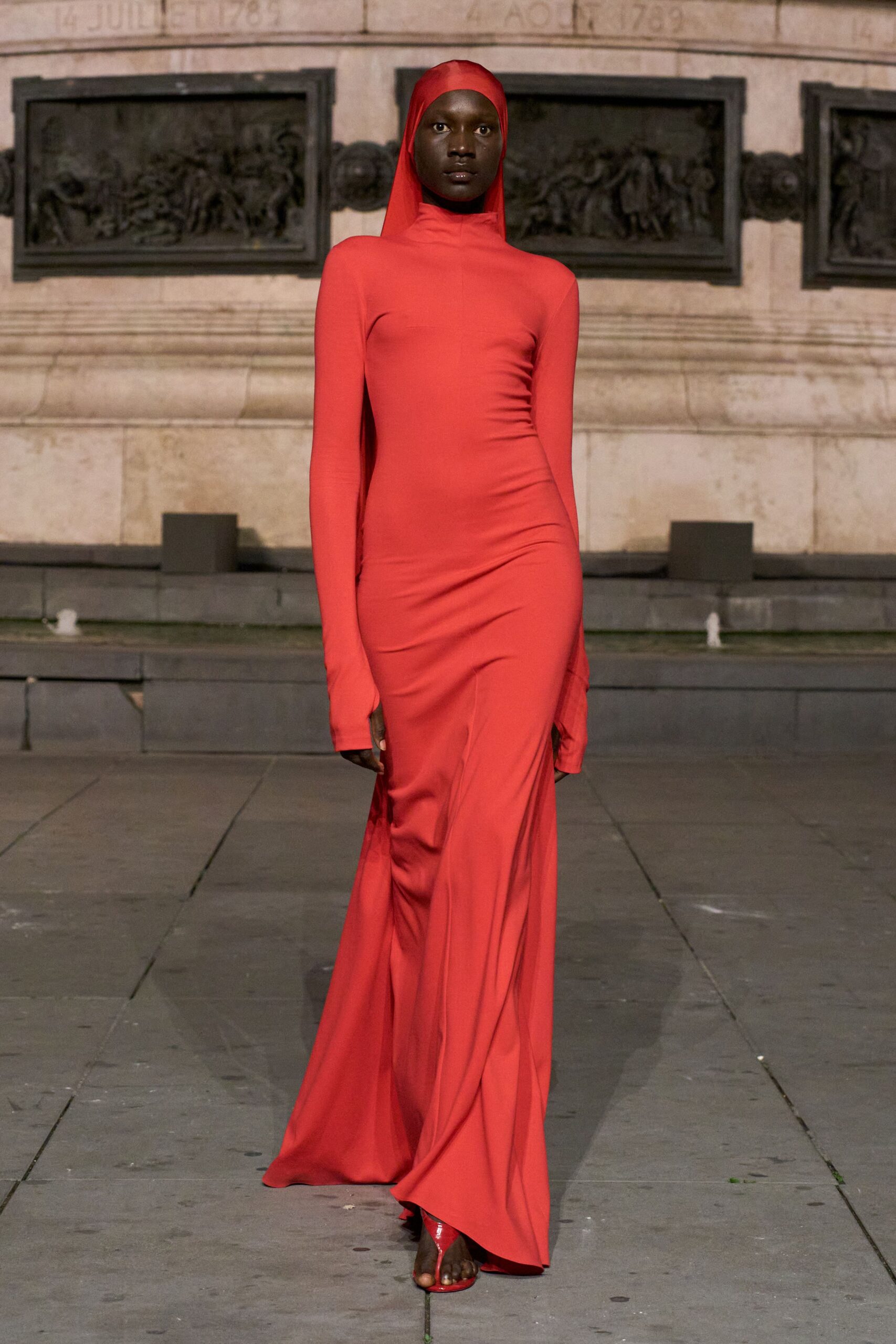 Tomato-red stretch jersey gown with built-in hood and a throat-high neck. Glossy red thong sandals peeking from the pooling hem. Crossing a public square just after rain, monument stone still giving back heat.