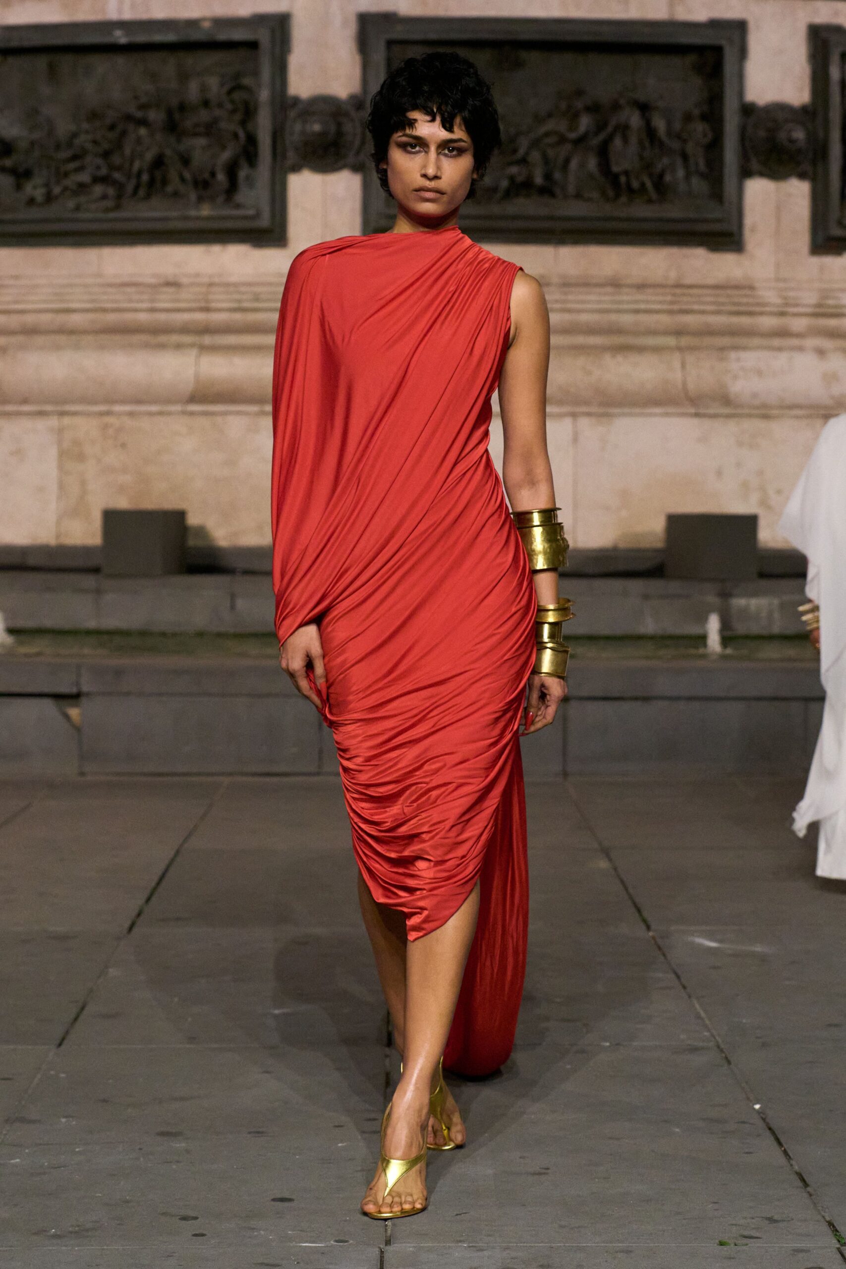 Tomato red jersey dress, one sleeve draped into tight ruching at the thigh. Stacked hammered brass cuffs climbing one forearm. Gold thong sandals with a narrow curved strap. Crossing a stone courtyard just before the wedding guests spill outside.