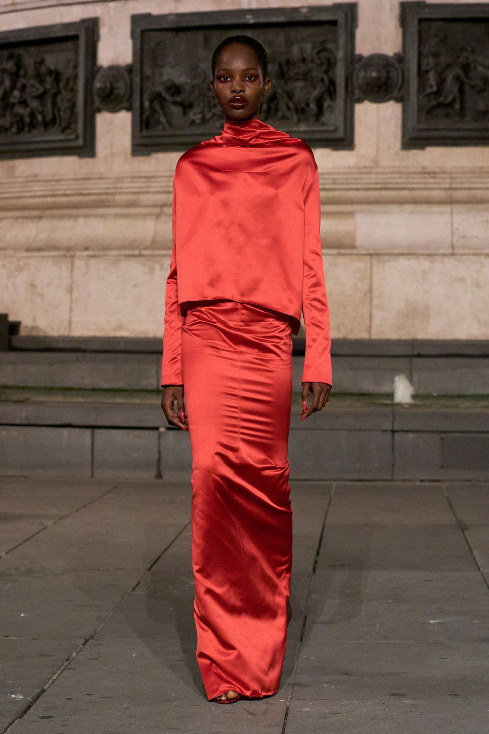 Tomato satin top draped into a soft funnel neck, hem cut boxy. Matching satin column skirt, ruched low at the hips and pooling. Red open-toe sandals barely showing under the hem. Crossing a stone square just after rain, headed somewhere with chandeliers.
