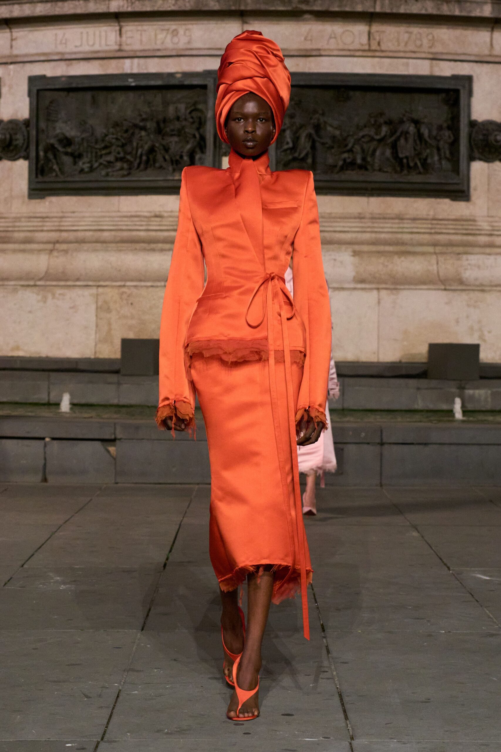 Towering tangerine satin headwrap, twisted into stacked folds. Sharp-shouldered satin jacket with a high neck and frayed hem. Column skirt in matching satin, raw edge brushing the ankles. Tangerine thong heels. Crossing the square just after rain, war memorial still holding the heat.