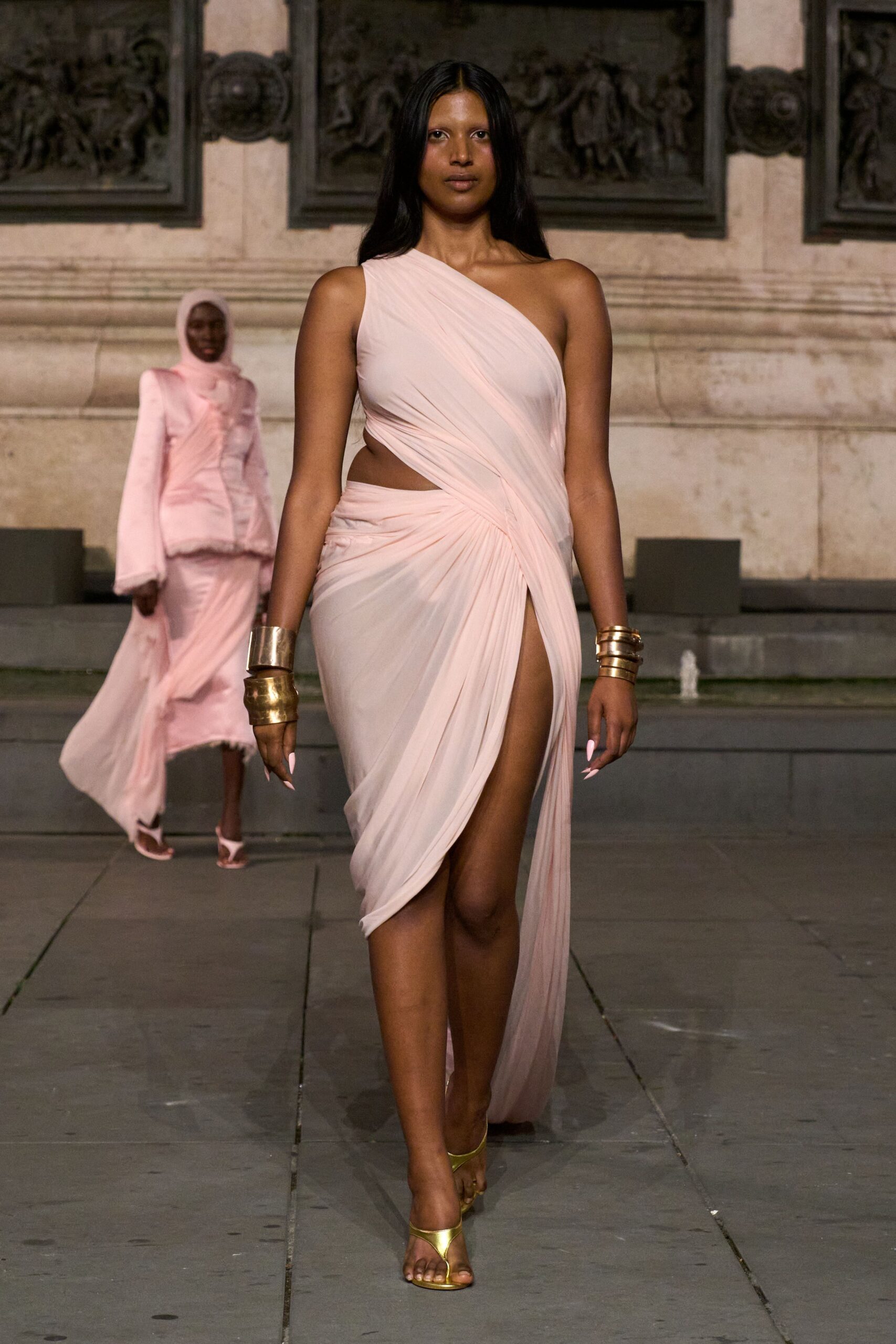 Blush one-shoulder draped dress in fine pleated chiffon, knotted low into a thigh slit. Stacked hammered gold cuffs on both wrists. Gold thong sandals with a narrow metallic strap. Crossing a stone courtyard just after the fountain switches on.