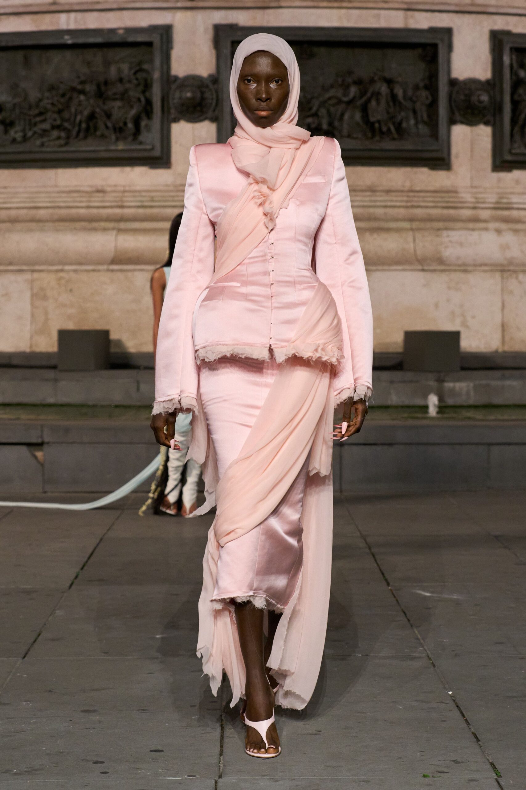 Blush chiffon headscarf wrapped close, one end draped at the throat. Pale pink satin dress coat, corseted through the waist with frayed hems. Sheer georgette panel spiraling diagonally across the body and skirt. Blush thong heels. Crossing a stone square just after a civil ceremony let out.