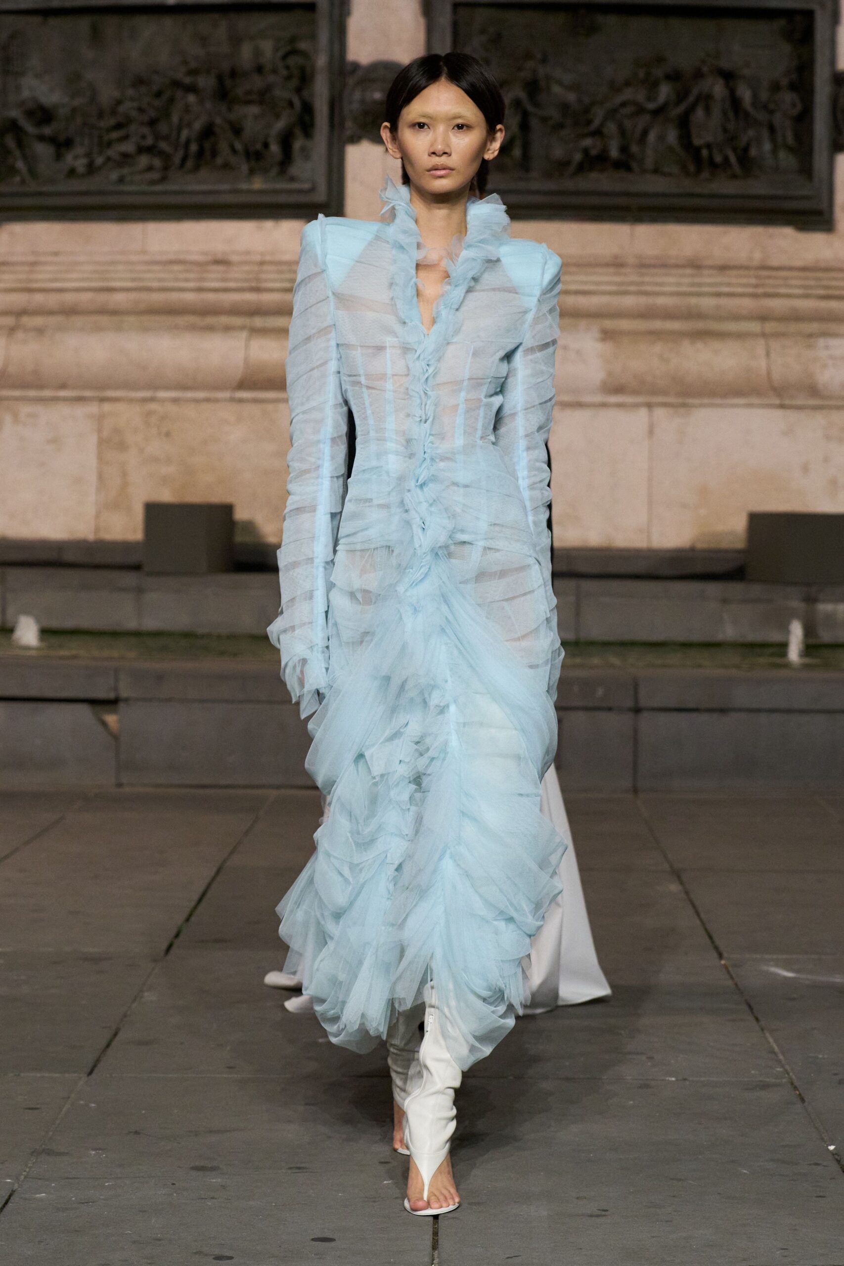 Powder-blue tulle gown, sheer striped panels and ruffles climbing the plunging front. White thong mules with wrapped ankles and a narrow pointed toe. Crossing a stone courtyard after the vows, veil already in one hand.