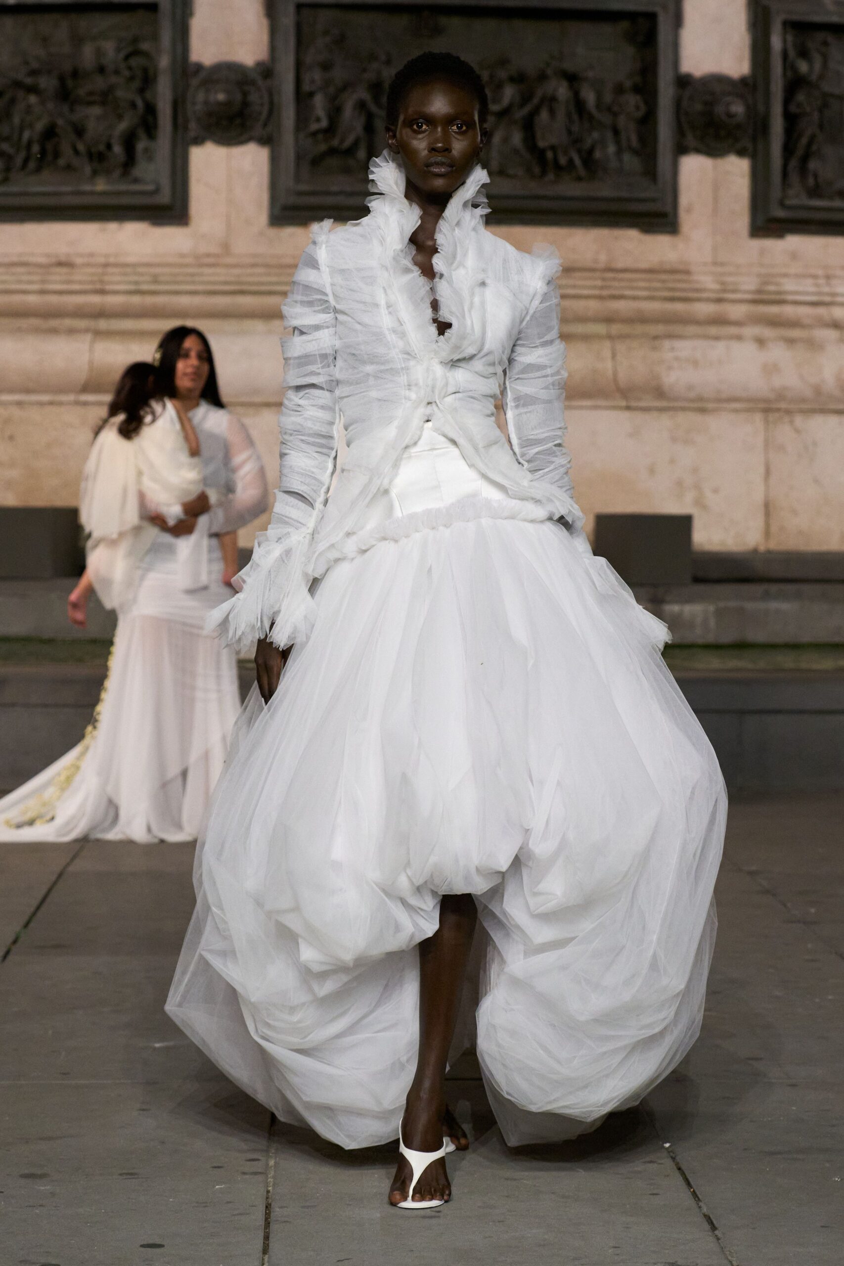 White sheer tulle blouse, ruched sleeves and a high frilled throat. Voluminous white tulle skirt, ballooned into deep stuffed folds at the hem. White thong sandals. Crossing a stone courtyard after the ceremony, dress still full of air.