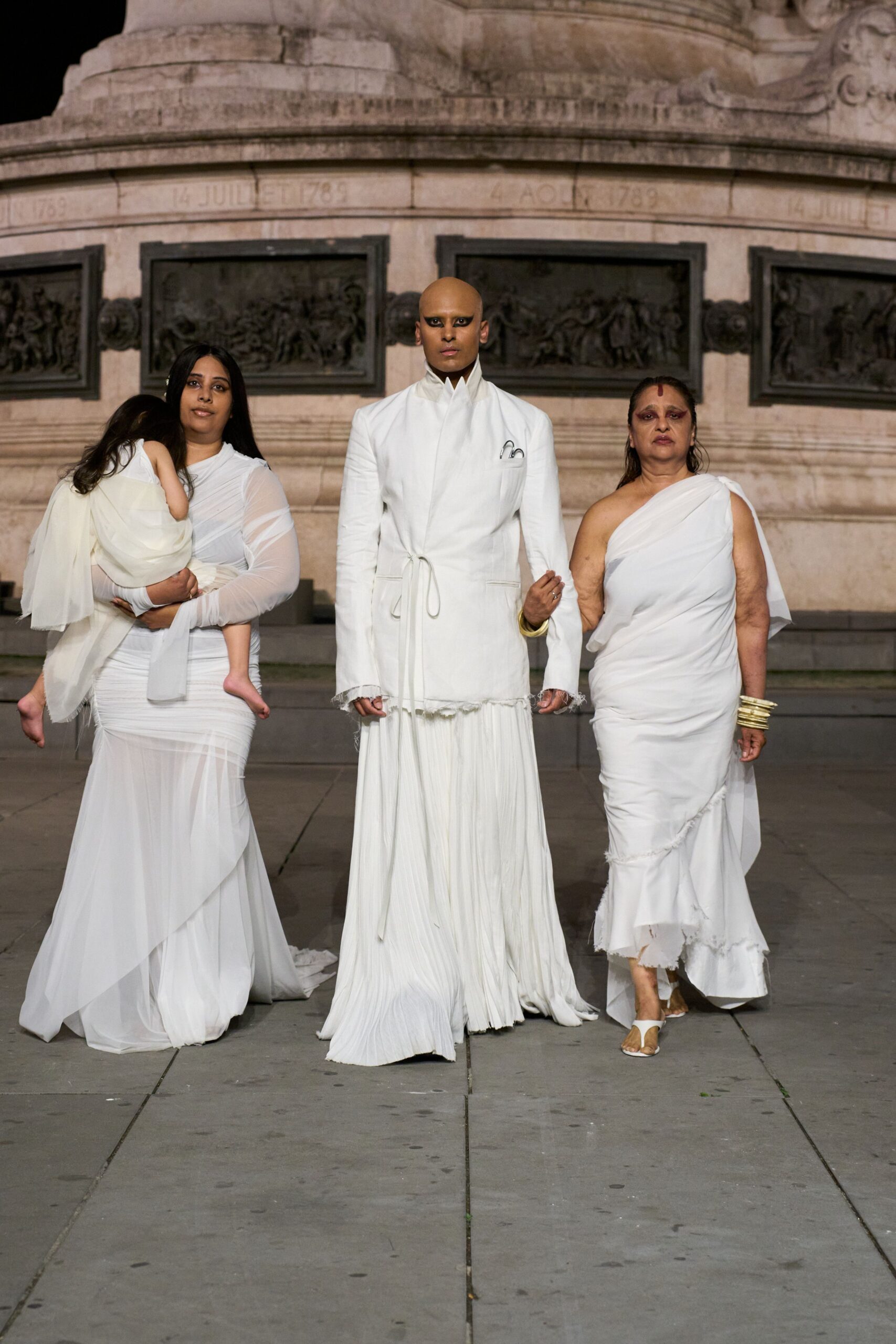 White longline jacket with a razor stand collar and frayed hem. Two silver safety pins tucked at the chest pocket. Floor-length white skirt in tight vertical pleats. Single gold bangle at the wrist. Crossing a stone square after a ceremony nobody can quite name.