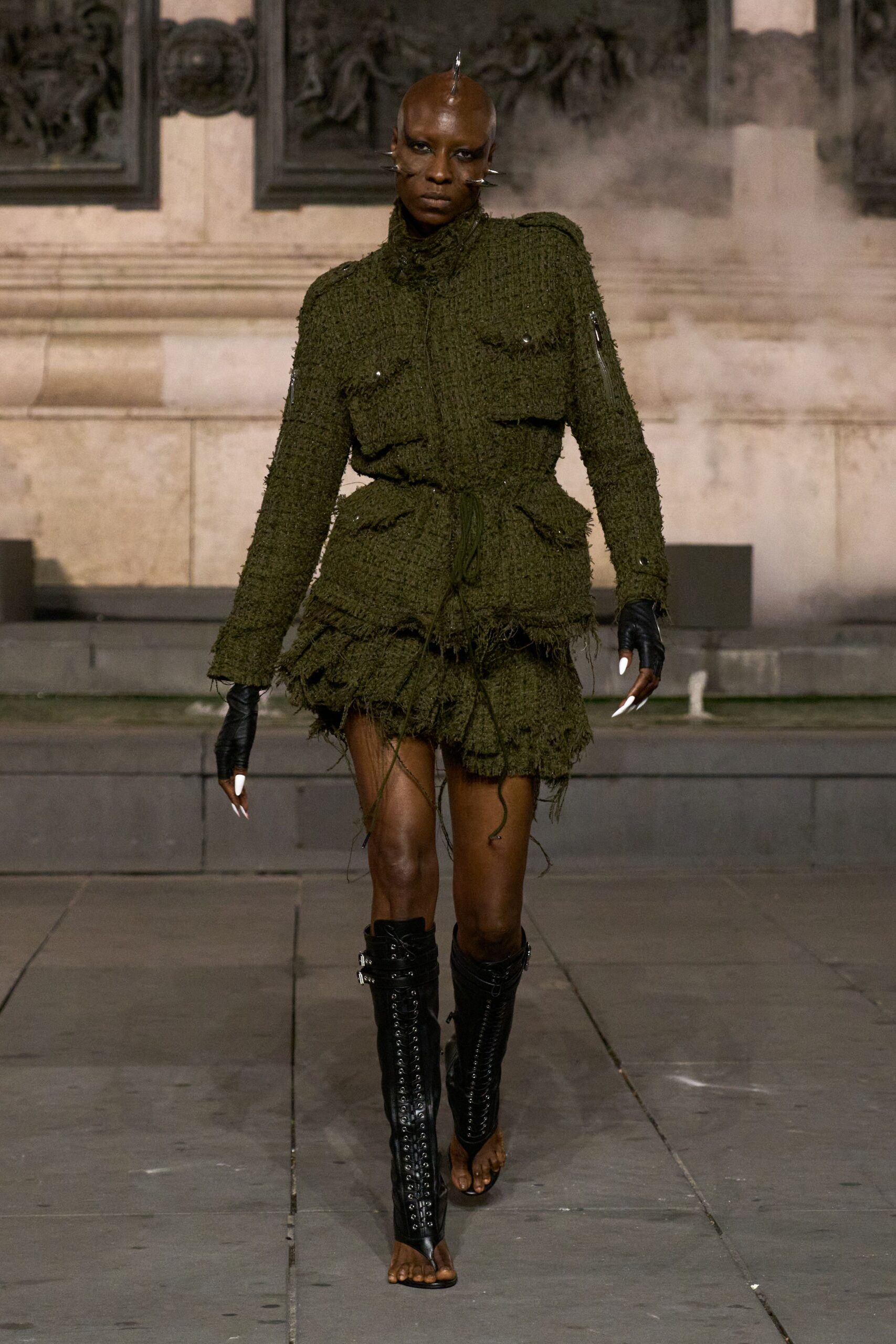 Metal spikes piercing the scalp line and both cheeks. Olive bouclé field jacket, frayed all over with cord-tied waist. Matching mini skirt built in ragged tiered flounces. Black fingerless leather gloves. Open-toe black leather boots, tightly laced up the shins. Whatever you wear home when the square is still smoking.