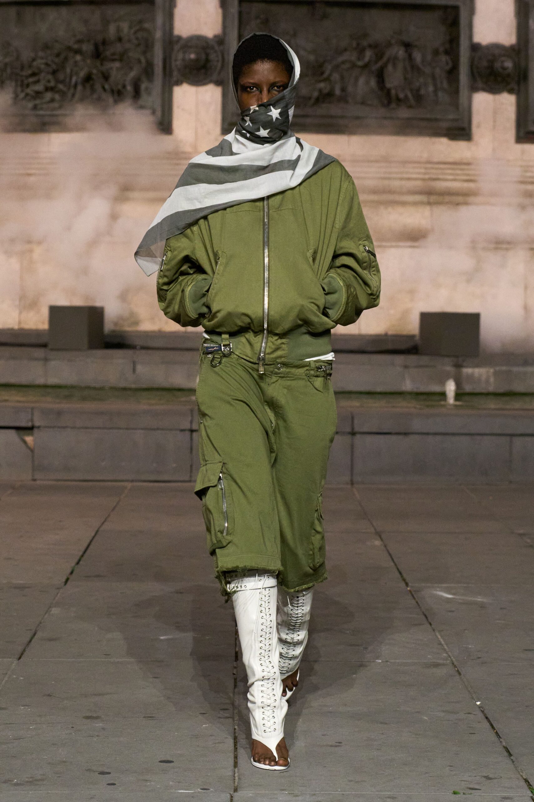 Oversized grey-and-white scarf, stars wrapped across the mouth. Olive flight jacket with a bright silver zip and sleeve pocket. Matching olive cargo skirt, D-ring tabs and a frayed raw hem. White lace-up stiletto boots, open at the toe. Crossing a monument square after the smoke machines have finally stopped.