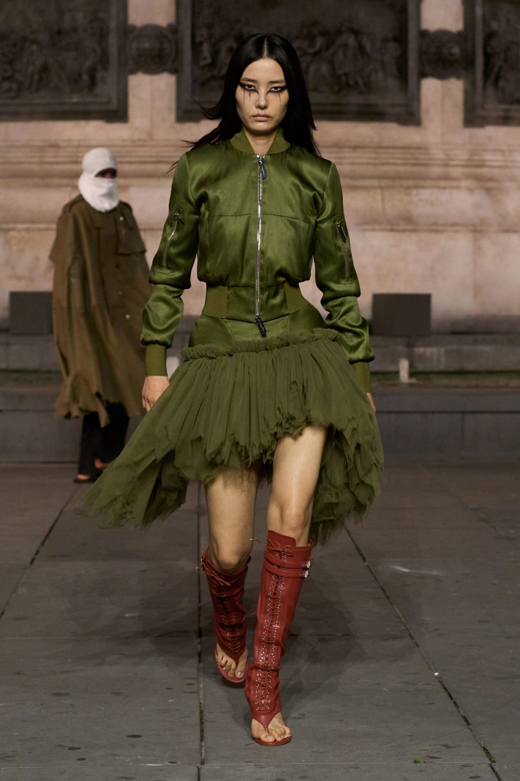 Olive satin bomber with sleeve utility zips and a dark zip pull. Asymmetric olive tulle skirt, ruched at the waist and ragged at the hem. Rust leather knee boots, front-laced and sliced open at the toes. Crossing a stone courtyard just before the storm finally breaks.