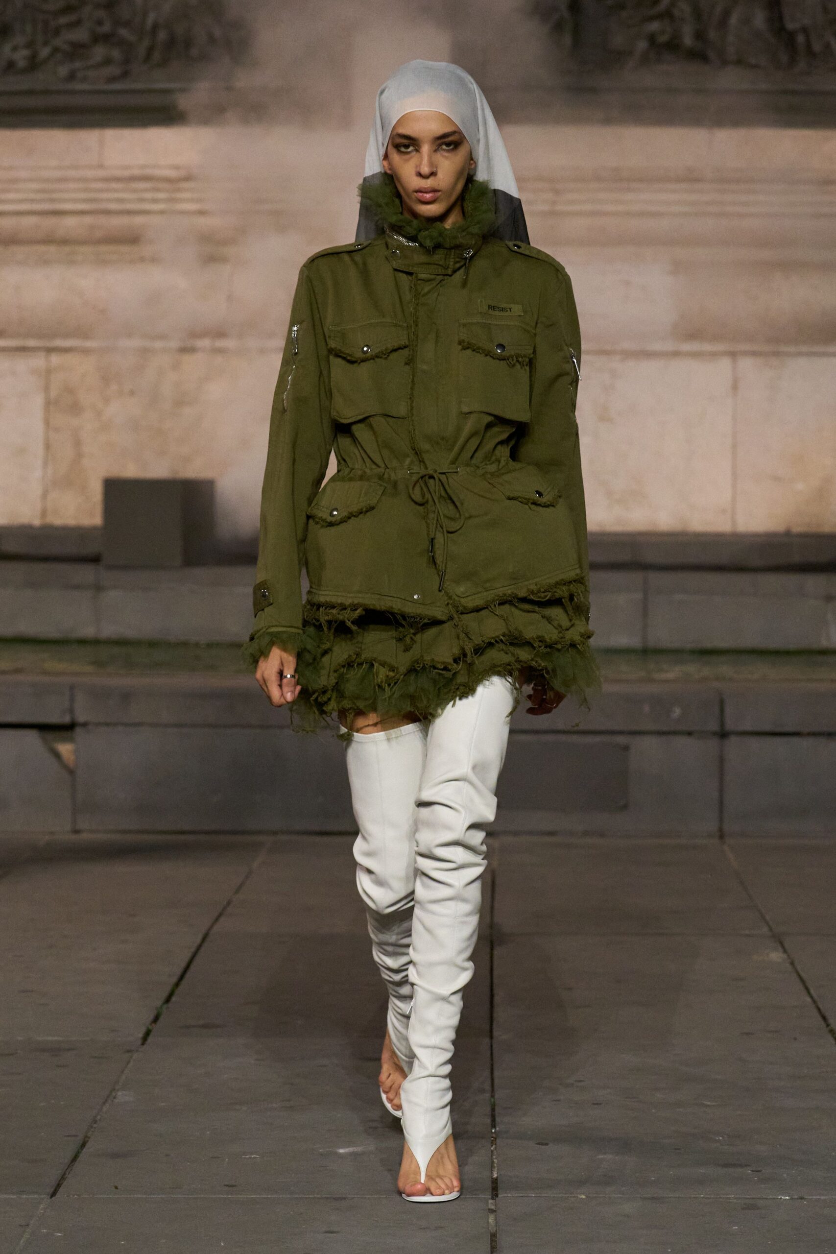 White veil falling to a black dipped back panel. Olive field parka cinched with drawcord, pocket flaps and hem left fraying. Layered tulle ruffle peeking from under the torn parka hem. White stretch thigh-highs cut into thong sandal fronts. Crossing a memorial plaza while the fountain throws cold mist.