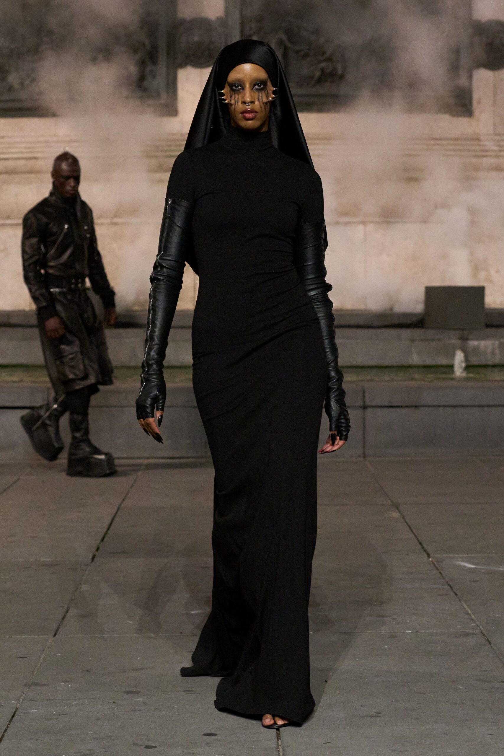 Black satin veil falling straight from a close skullcap. Column turtleneck gown in matte jersey, slightly ruched through the hip. Black leather opera gloves, zipped at the upper arm. Bone-colored claw pieces hooked along both cheeks. Small silver septum ring. Crossing a monastery courtyard while the incense still hangs low.