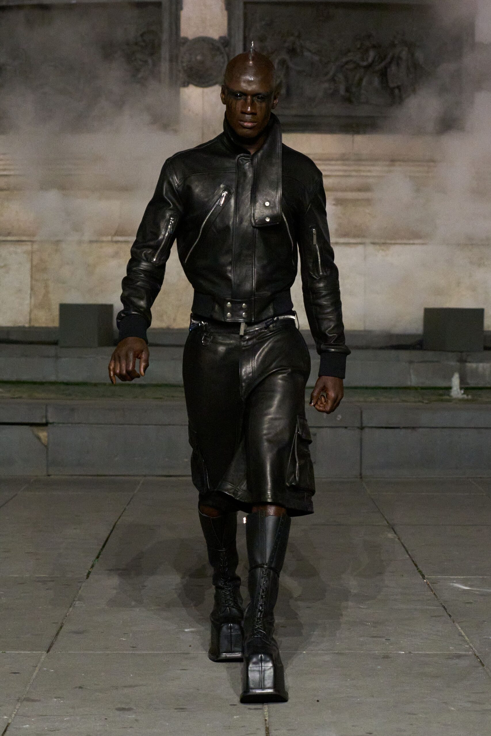 Black leather flight jacket with zippered sleeves and a long snap-tab collar. Black leather kilted shorts with cargo pockets and a ring at the waistband. Tall black lace-up platform boots, one shin capped in glossy armor. Crossing a stone courtyard while the fountains are still running in fog.