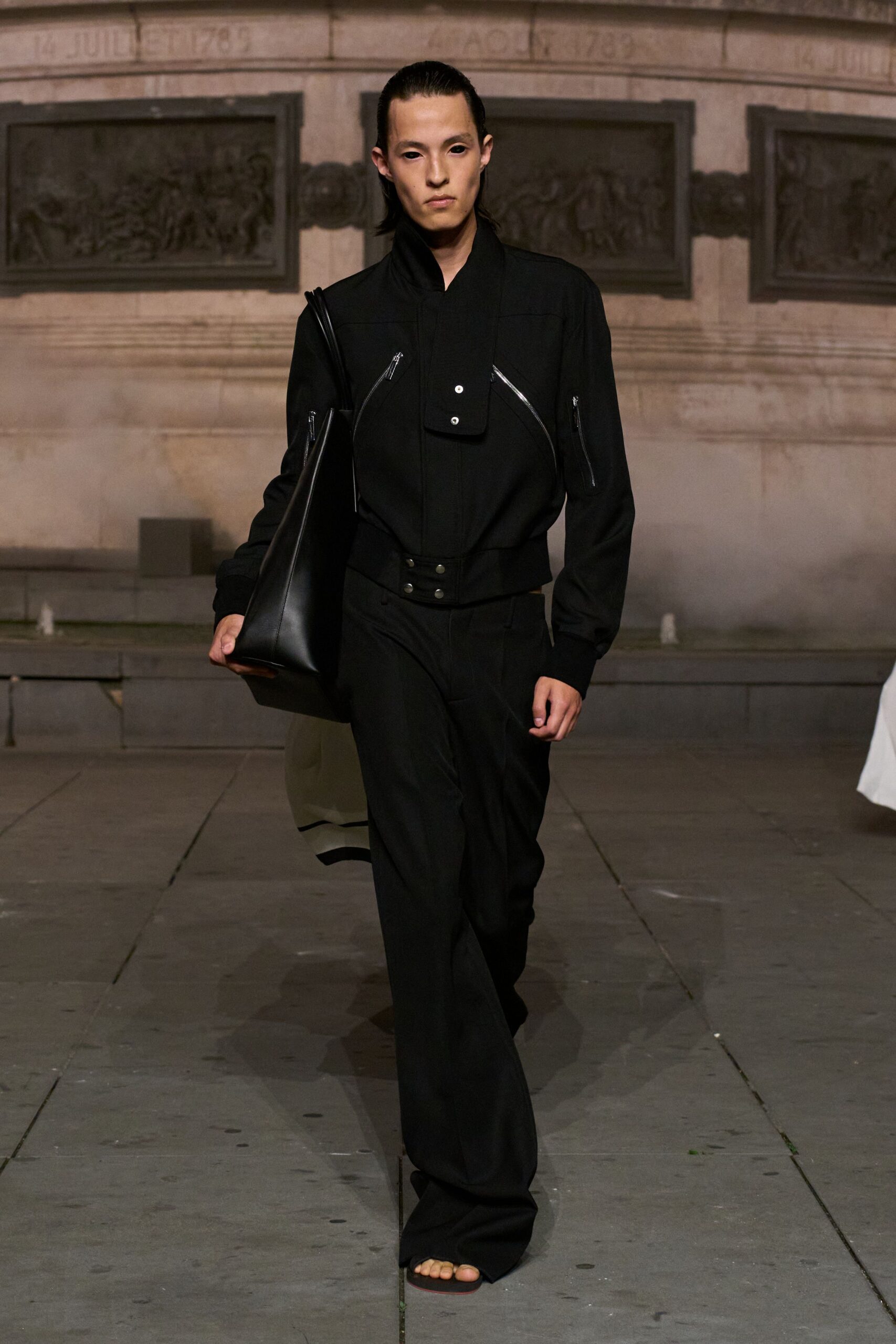 Black jacket with a funnel neck, curved silver zips and a bib placket. Oversized black leather tote tucked hard under one arm. Black trousers falling long over the foot, held by a double-snap waistband. Barely-there thong sandals exposing the toes. Crossing a stone square after rain, keys somewhere at the bottom of the bag.