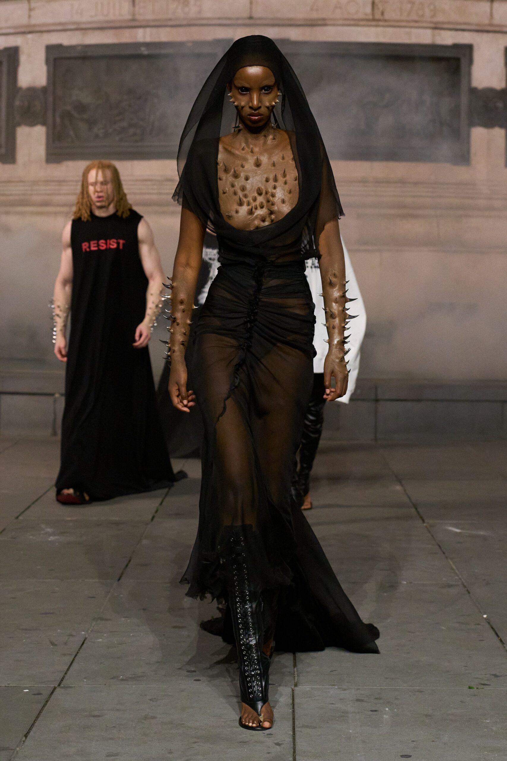 Black chiffon veil falling from a close skullcap. Sheer black jersey gown, deeply cowl-necked and ruched up the center. Nude prosthetic spikes running from shoulders to wrists. Black lace-up shin guards ending in bare-toe sandals. Crossing a stone square after midnight, church bells still shaking the air.