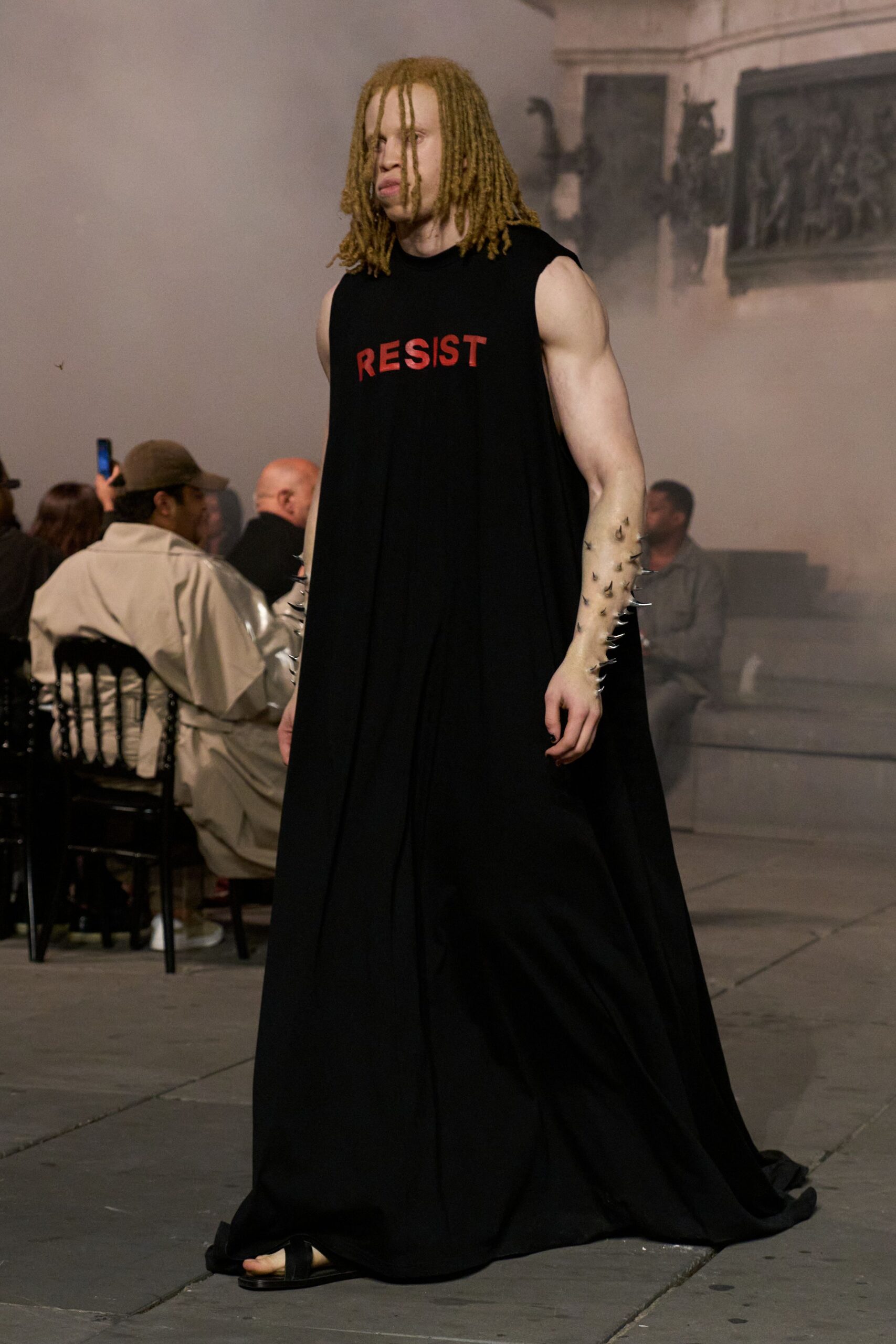 Black sleeveless jersey column, red RESIST letters stark across the chest. Clear forearm pieces studded with silver thorn-like spikes. Black slide sandals, bare toes visible beneath the dragging hem. For reading a manifesto in a stone hall gone smoky with incense.