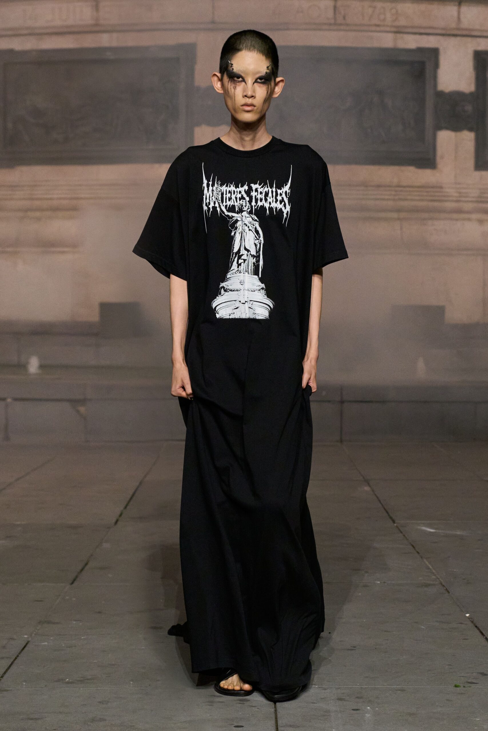 Oversized black cotton T-shirt with a cracked white monument print. Floor-length black skirt in heavy jersey, bunched up in both hands. Flat black thong sandals barely visible under the dragging hem. Crossing a stone square after midnight, fountain switched off for cleaning.