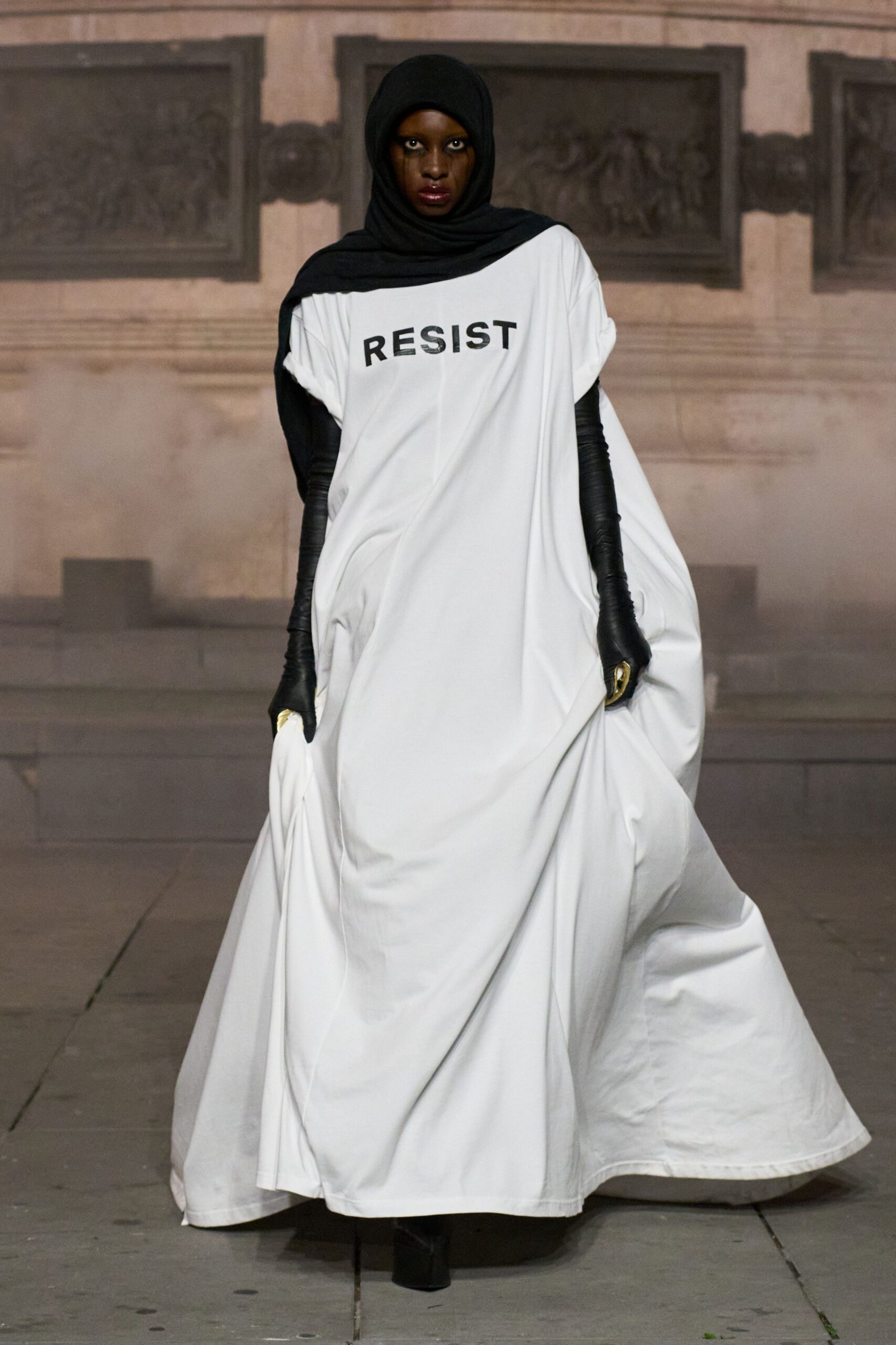 Black jersey wrapped close, one end laid broad across the shoulder. White floor-length jersey caftan with RESIST printed stark across the chest. Black opera gloves with small gold finger caps catching the light. Black boots barely visible under the dragging hem. Crossing a smoke-hazed civic square after the crowd has thinned.
