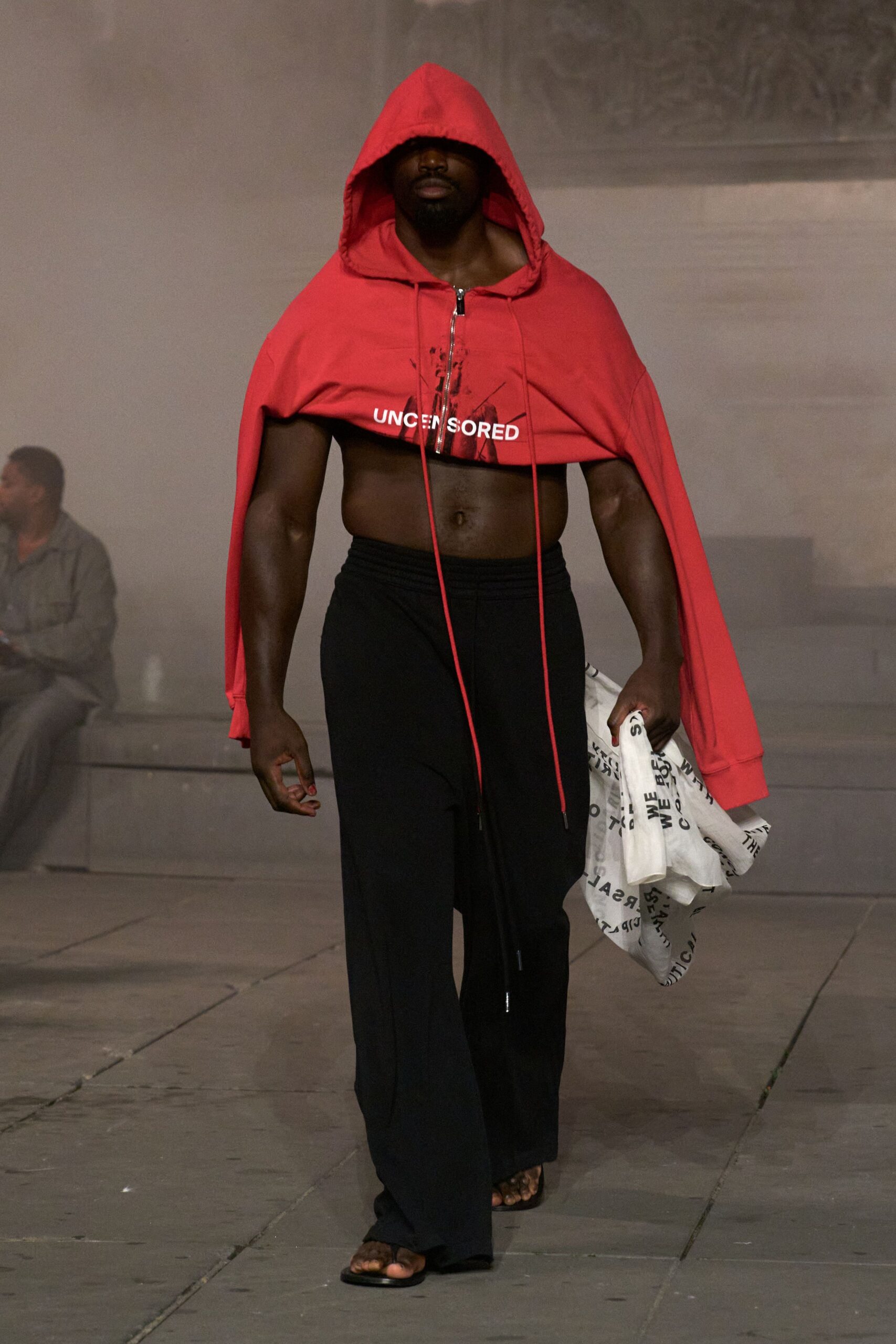 Tomato jersey hoodie chopped above the ribs, zip ending under printed text. Black fluid trousers with a densely shirred waistband and long rise. Flat black slides, toes fully exposed. White cloth tote crumpled in hand, scattered with black lettering. Leaving the community pool at dusk with protest flyers gone damp.