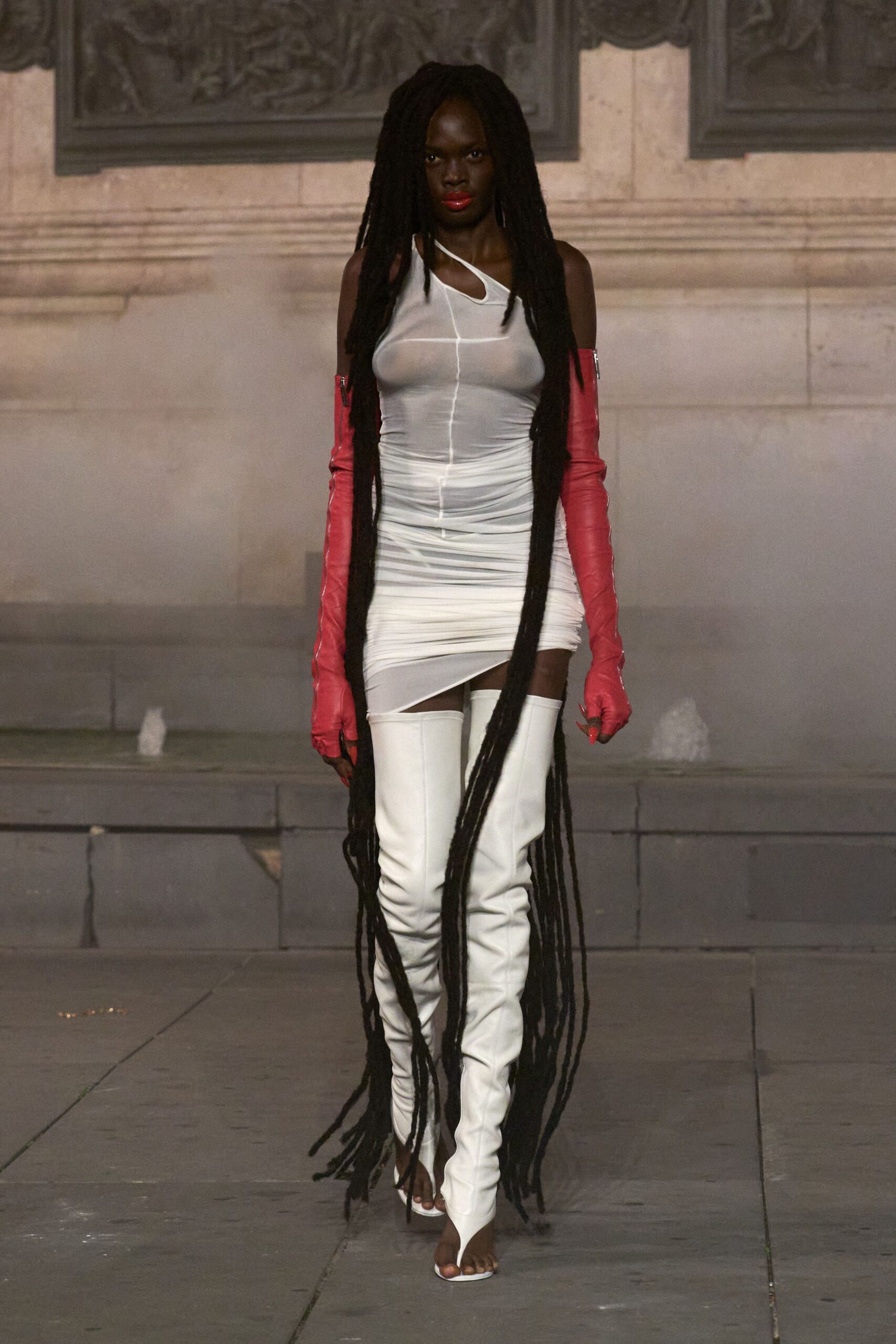 White stretch jersey dress, one-shoulder cutout neckline and ruched hips. Watermelon leather opera gloves, silver zips running the outer arm. White thigh-high boots with split-toe fronts and sharp cone heels. Crossing a stone courtyard while the fountain mist catches on your skin.