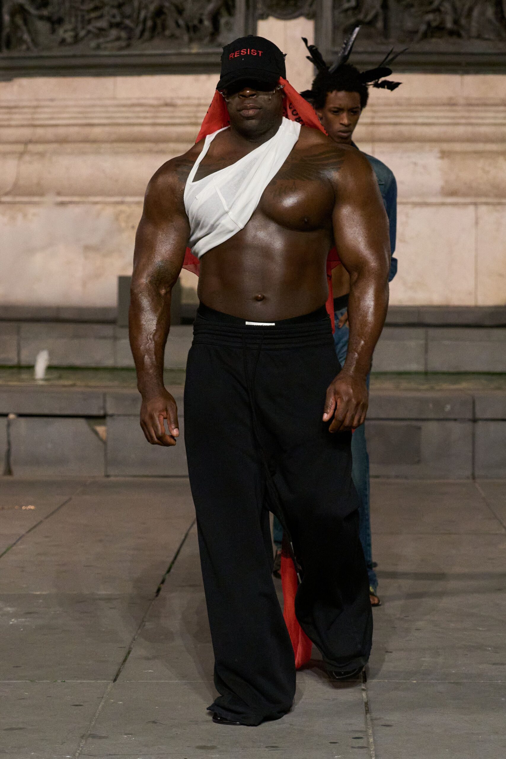 Black baseball cap with red lettering and a long scarlet veil. White jersey bandage top wrapped diagonally across one shoulder. Black wide-leg trousers with a ribbed waistband and pooled hem. Pointed black shoes peeking out. Crossing the square after a rally, fountain spray still on the pavement.