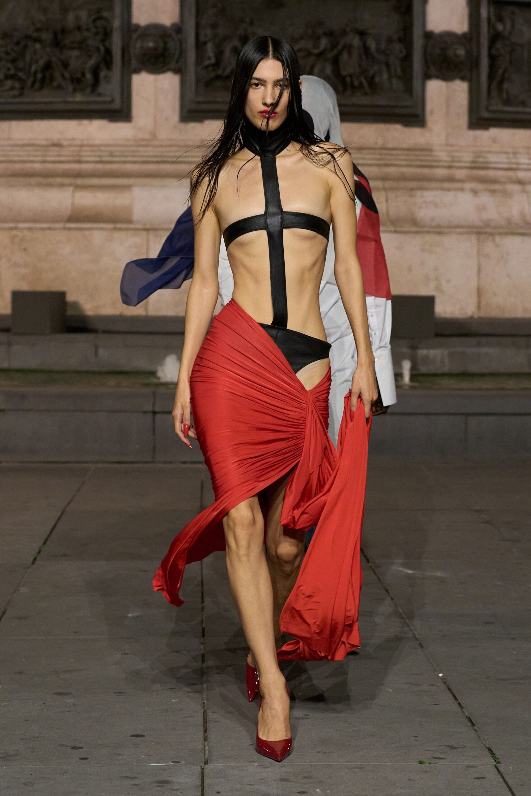 Black leather harness bodysuit with a high collar and stark center strap. Tomato red jersey wrap skirt, ruched hard at one hip into a trailing panel. Patent oxblood pointed pumps. Crossing a rain-dark plaza after midnight with the gala still roaring upstairs.