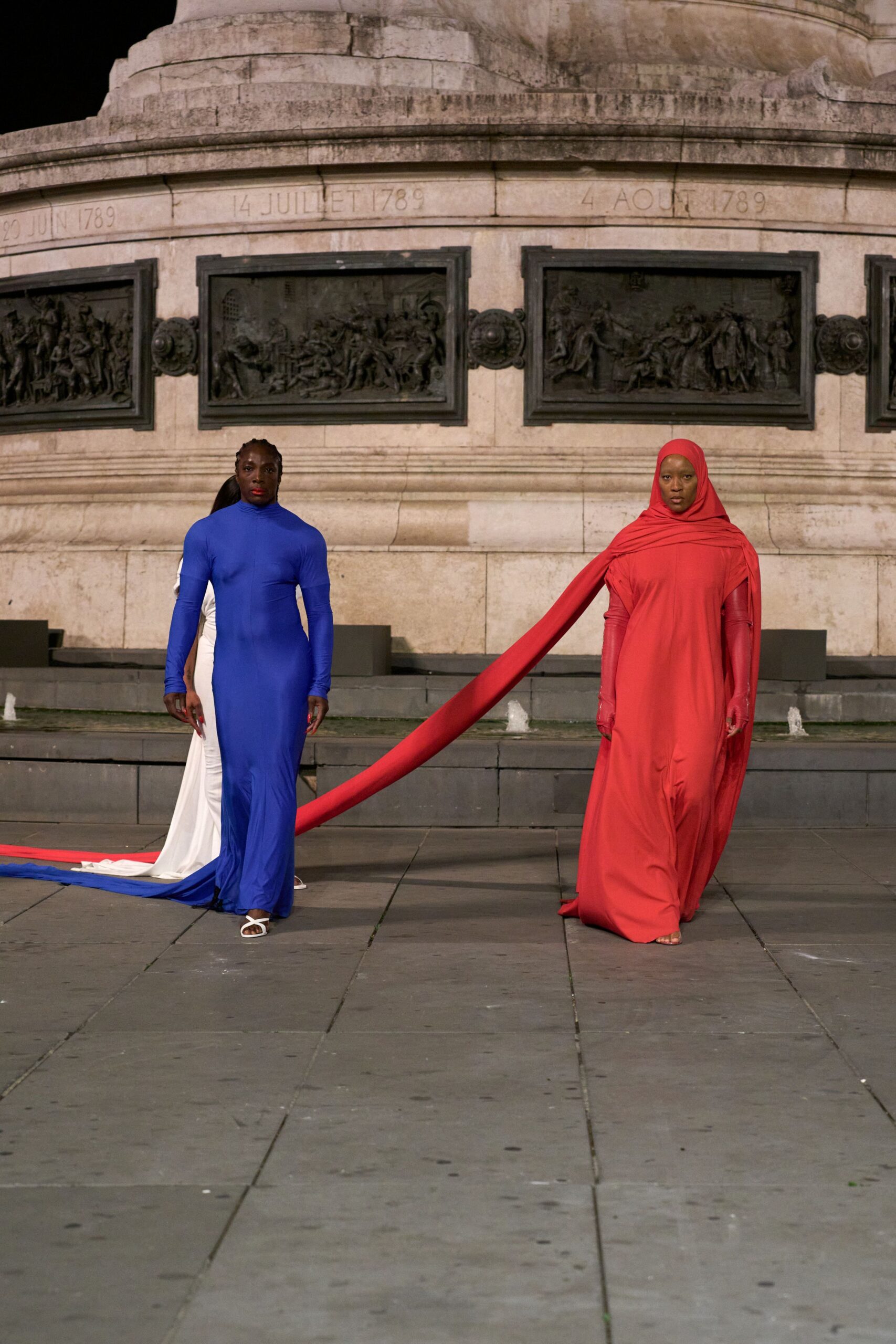 Tomato-red jersey column gown with a wrapped hood and floor trail. Matching opera gloves in sheer stretch mesh, seams running straight to the wrist. Barely visible nude sandals. Crossing a public square after dark while the monument keeps its heat.