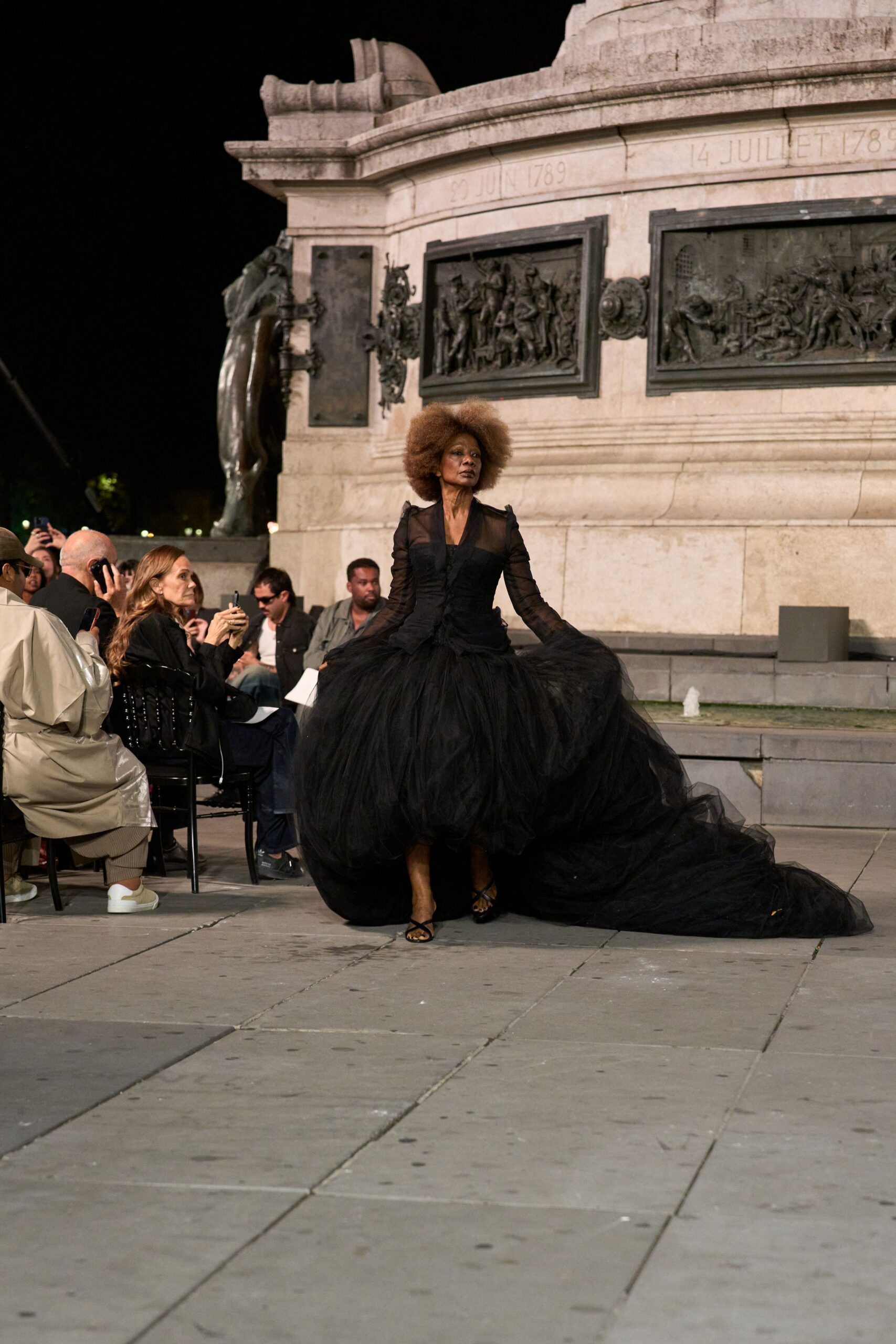 Black tulle gown with sheer sleeves, ruched bodice and vast trailing skirt. Barely there black straps crossing the toes. Crossing a public square after midnight, hem drinking up the paving dust.