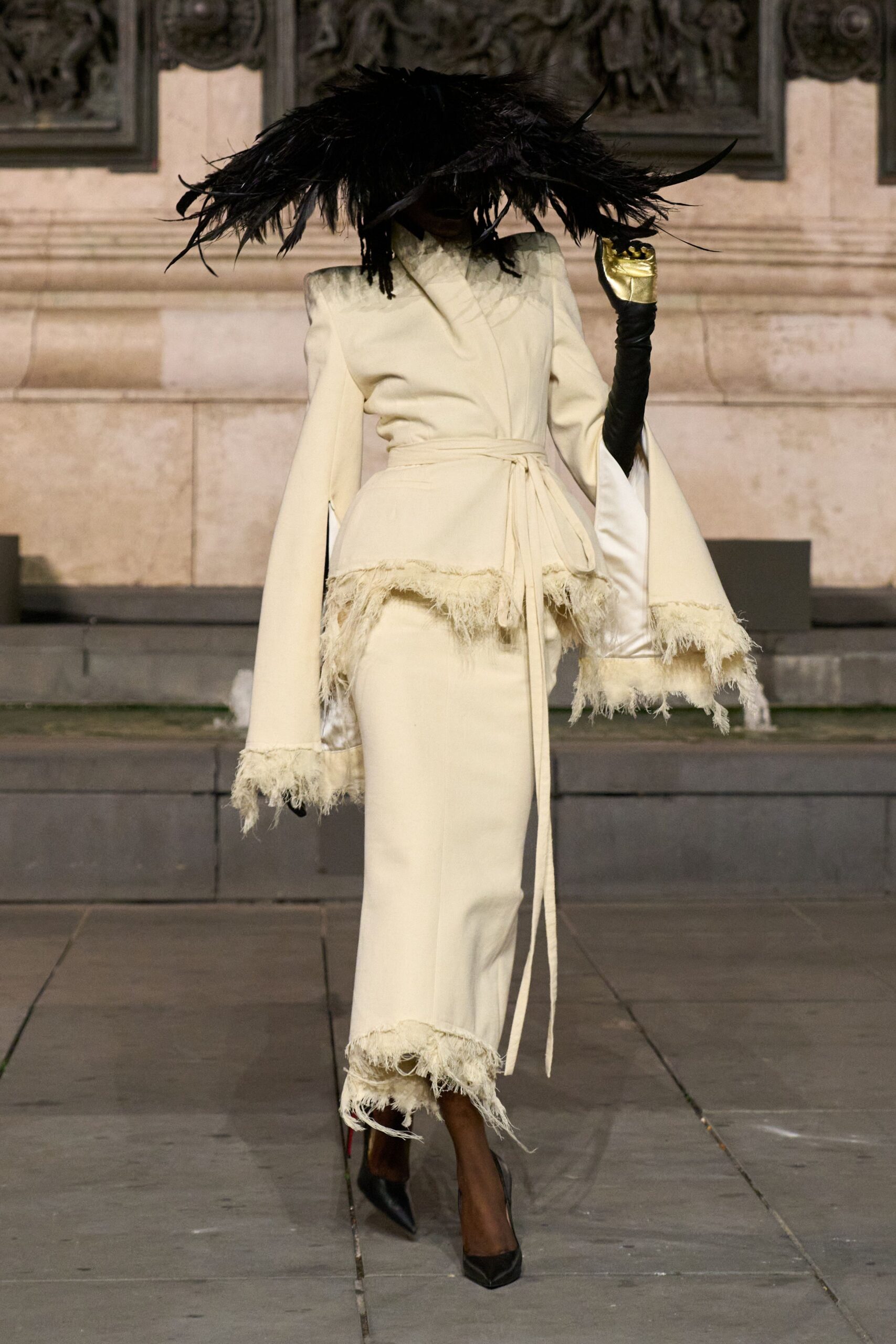 Huge black feather brim drooping low over the face. Ivory wrap jacket with slit sleeves and shaggy fringe at the hem. Opera-length black gloves, one capped with a pale gold cuff. Ivory straight-leg trousers with feathery fraying around the ankle. Black pointed pumps. Crossing a stone courtyard just before the bells start.
