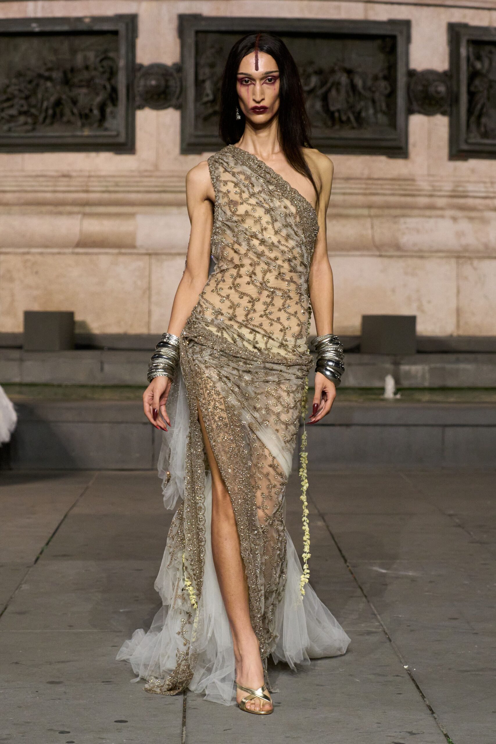 One-shoulder nude tulle gown densely beaded in pewter vinework, hip drape falling into a high slit. Stacks of hammered silver bangles on both wrists. Gold leather sandals with crossed straps at the toes. Stone courtyard after dusk, fountain noise catching in the beaded train.