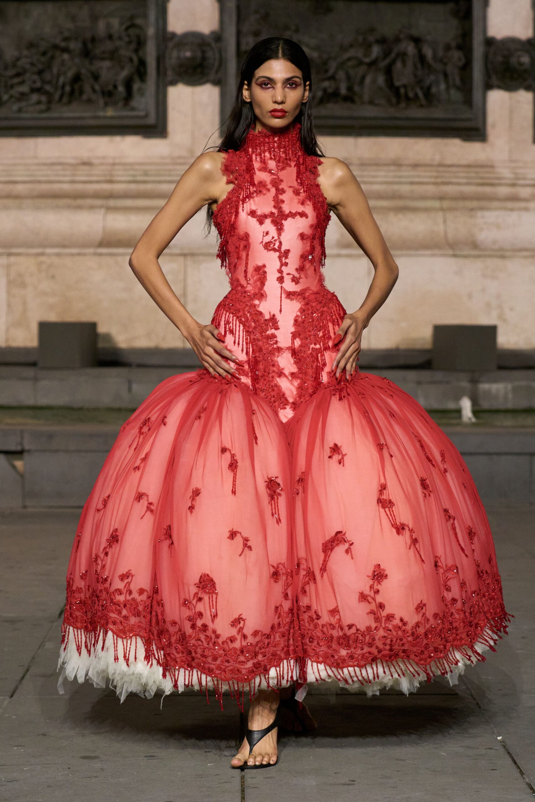 Blush ball gown with scarlet beaded appliqué, fringed high neck and pannier hips. Black thong sandals, nearly lost under the tulle hem. For arriving at a palace wedding after the orchestra has already started.