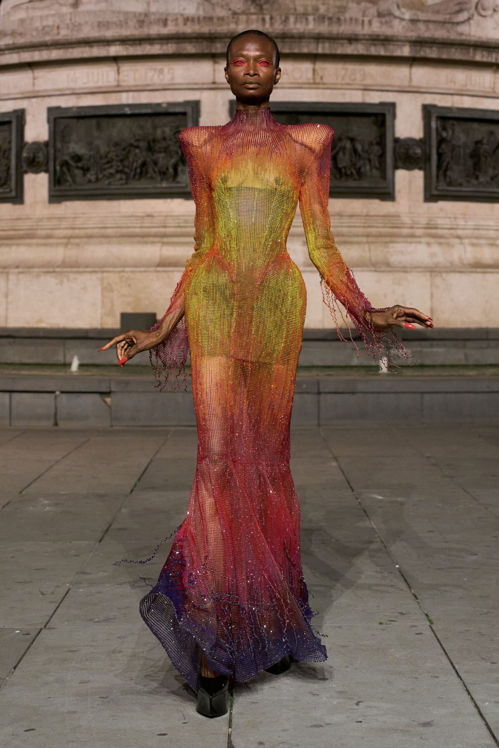 Transparent crystal mesh column fading from citron to ember to violet, beaded fringes dripping from the cuffs. Black patent square toes peeking beneath the pooled hem. Dawn in a stone square, fountain spray still hanging in the air.