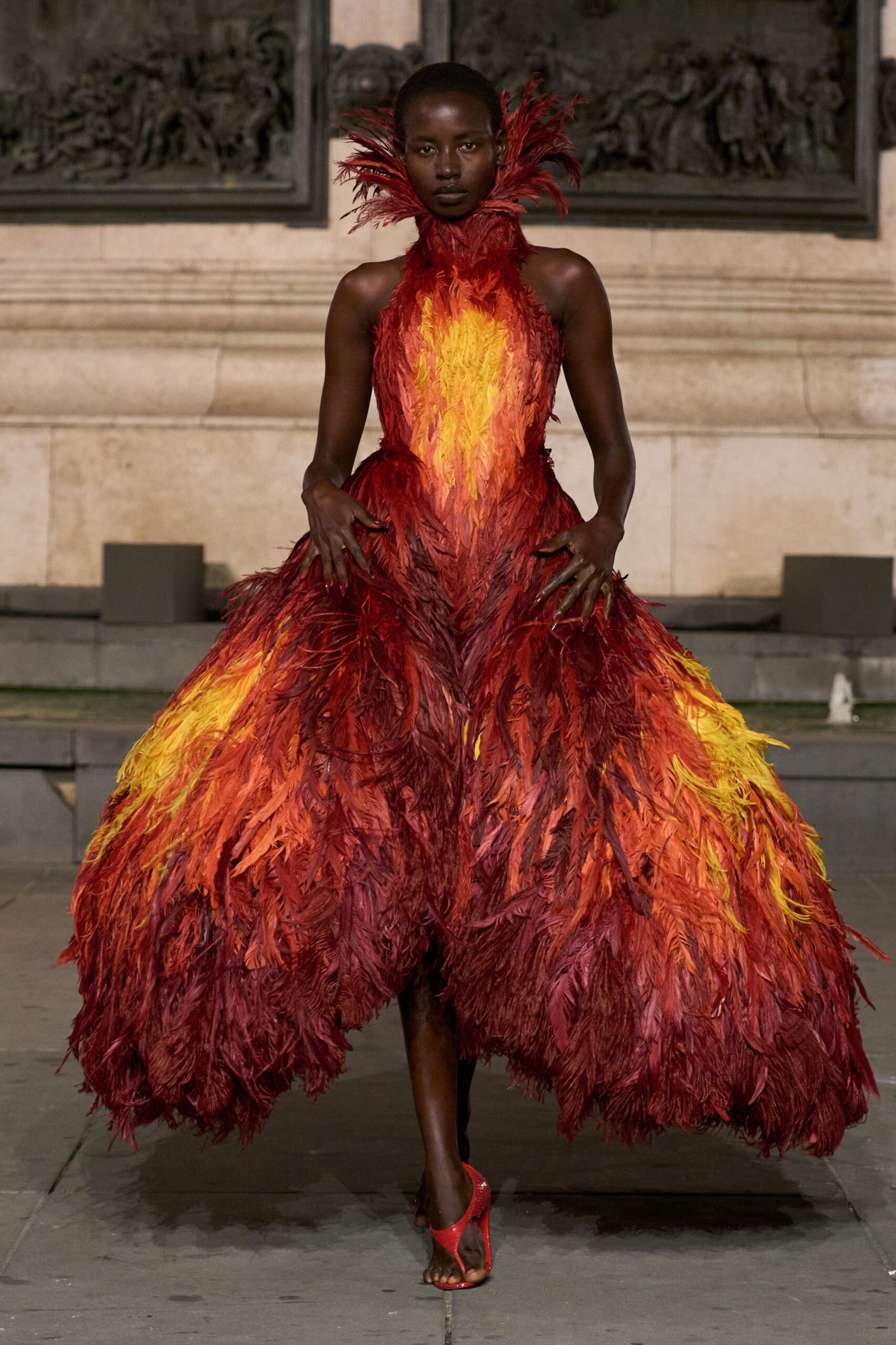 Strapless flame-plume gown, scarlet to marigold feathers swelling into panniered volume. Matching feather ruff shooting upward in singed-looking quills. Red thong heels with a glittered finish. Crossing a stone courtyard just after the fireworks ash starts to fall.