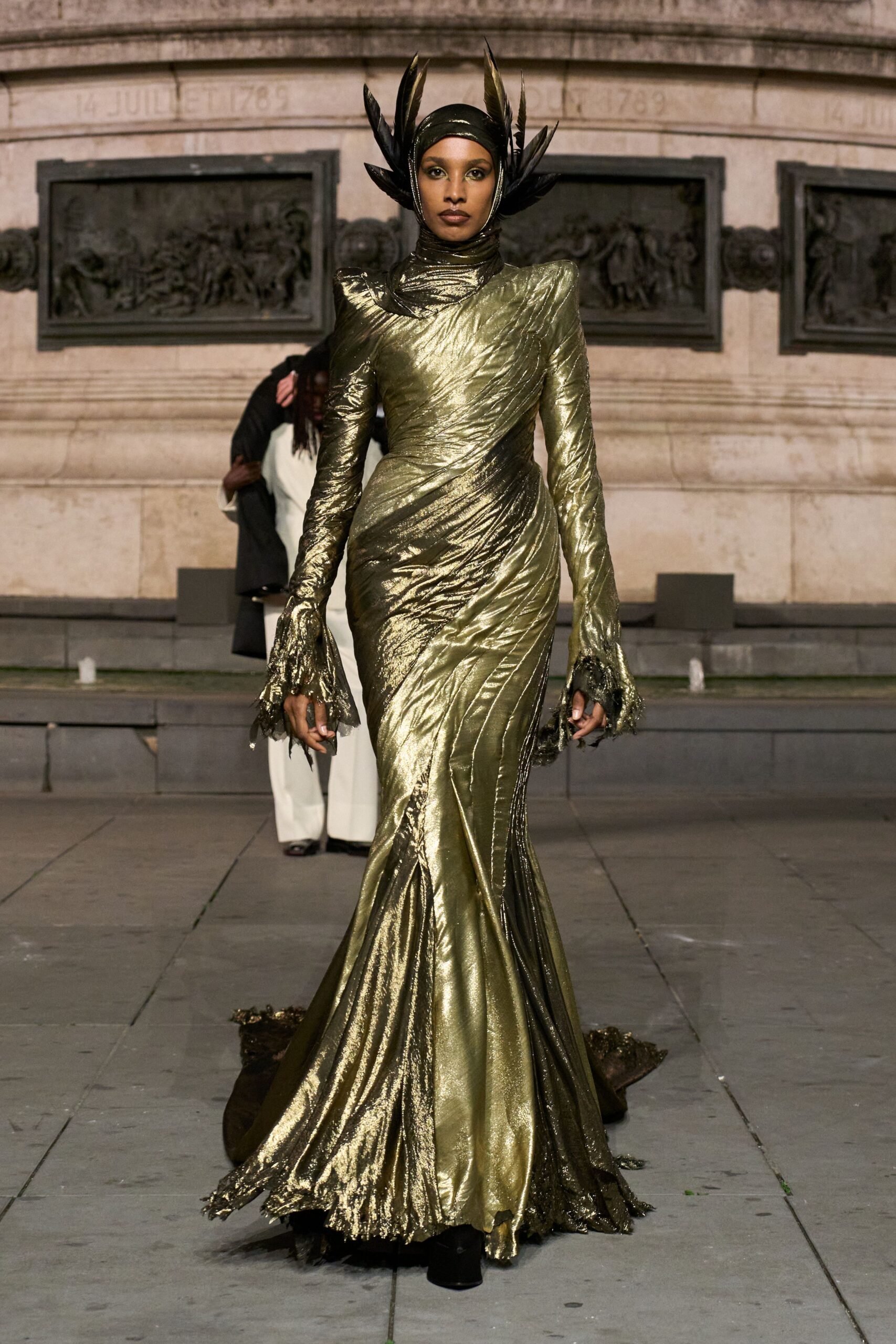 Black lacquered feathers flaring from a tight wrapped hood. Olive-gold lamé gown spiraled in ruched bands, cuffs and train in ragged black tulle. Black platform boots barely breaking the hem. At a monument after rain, when the pigeons have finally scattered.