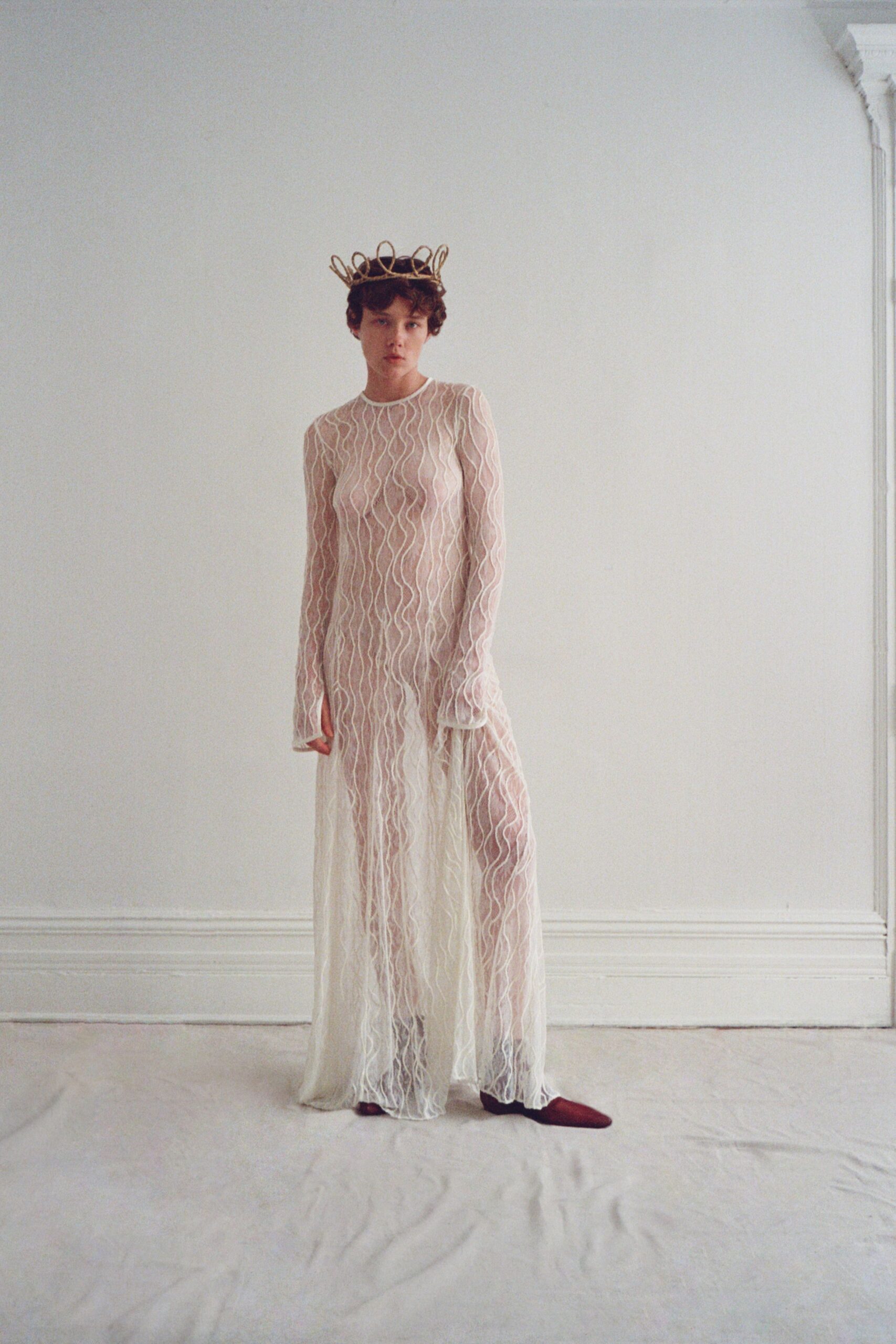 Looped wire crown perched lightly in the curls. Sheer ivory mesh column traced with raised squiggle embroidery. Opaque cream slip falling shorter beneath the transparent hem. Burgundy pointed flats peeking out under the pooling edge. Morning in a drafty townhouse, still wearing the pageant prize.