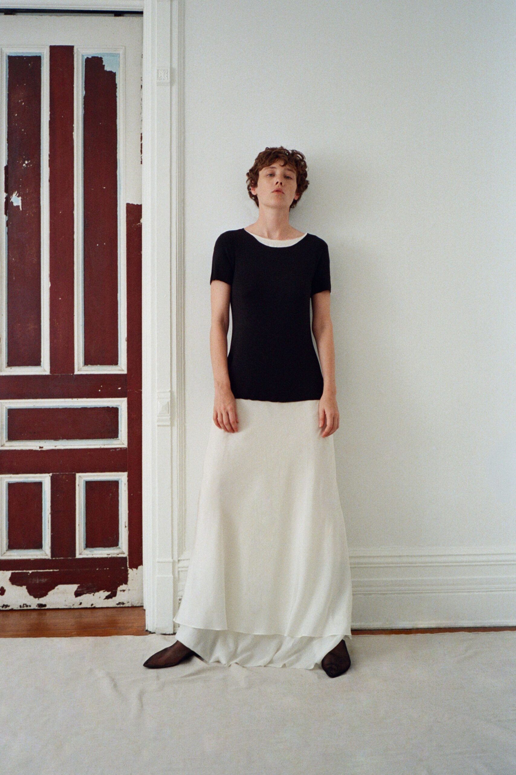 Black knit top with a white bateau inset skimming the collarbone. Ivory bias-cut skirt in matte fluid cloth, pooling over a second hem. Dark mesh flats, squared off at the toe. In a rented apartment after the sheets are off the furniture.