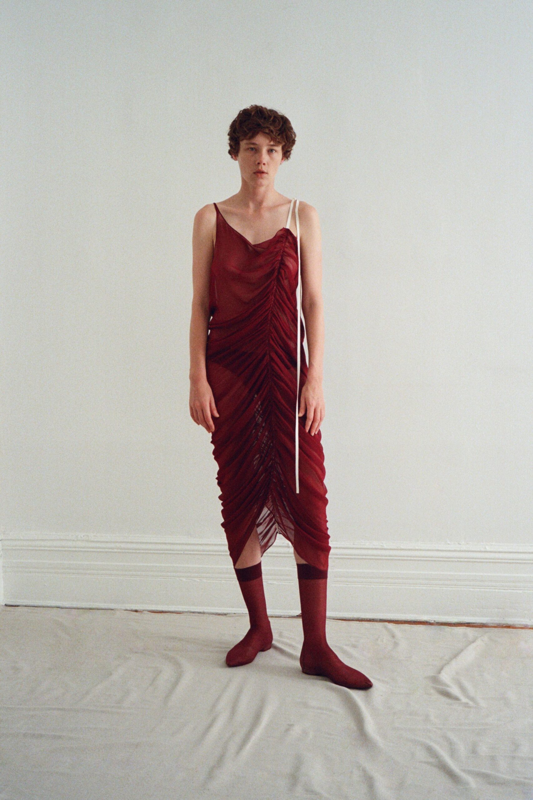 Oxblood slip dress in sheer mesh, ruched hard down the center seam. Long ivory cord strap hanging past the knee from one shoulder. Matching oxblood socks with a darker band circling the calf. The hour before a small concert, standing barefoot on dust-sheeted floorboards.