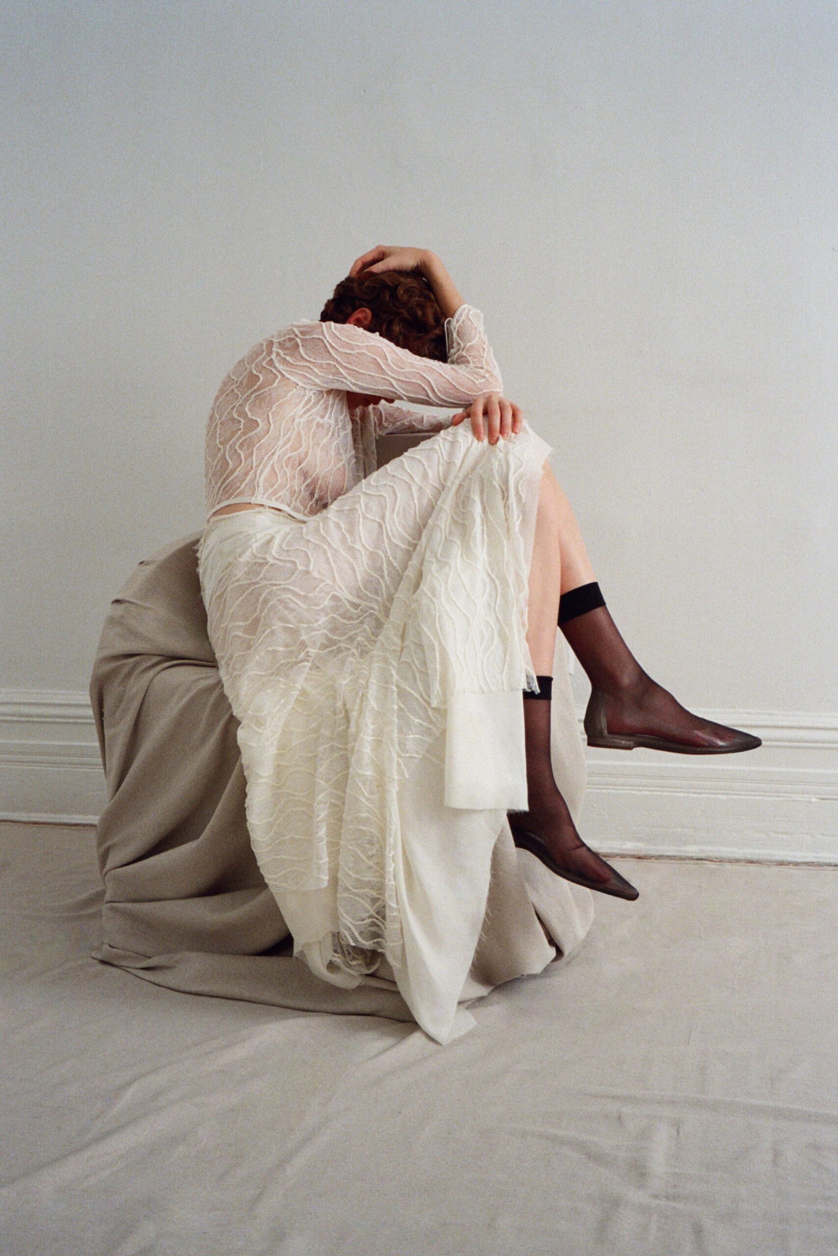 Sheer ivory mesh top traced with raised squiggle embroidery. Matching ivory skirt over a denser underlayer, raw hem grazing the floor. Black sheer ankle socks with opaque bands. Pointed brown leather flats, vamp cut low. Sitting on the sheet-draped chair after the registry office, nobody speaking first.