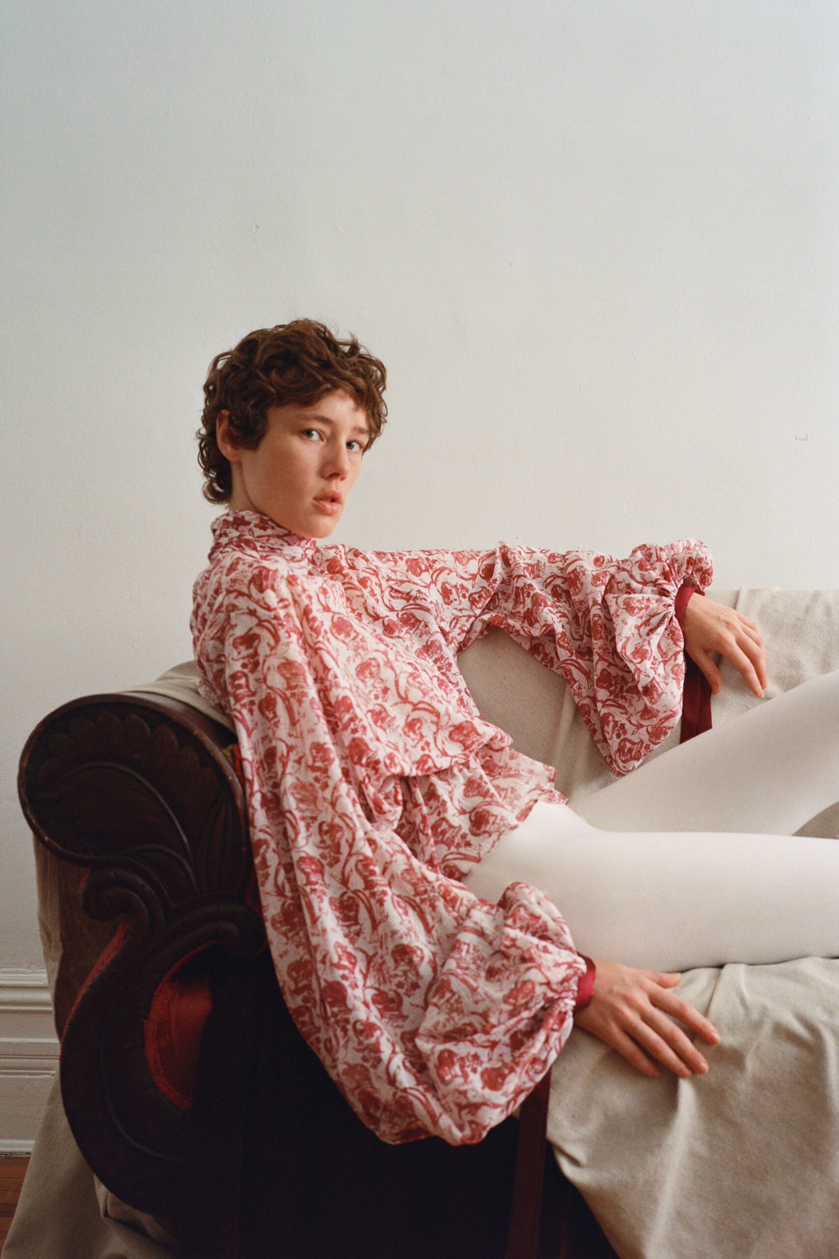 Rose toile blouse with a high frilled neck and gathered bishop sleeves. Opaque white tights, unbroken from hip to pointed toe. Wine satin inner cuffs peeking past the ruffled sleeve ends. Afternoon in a rented apartment, still in costume after opening the curtains.