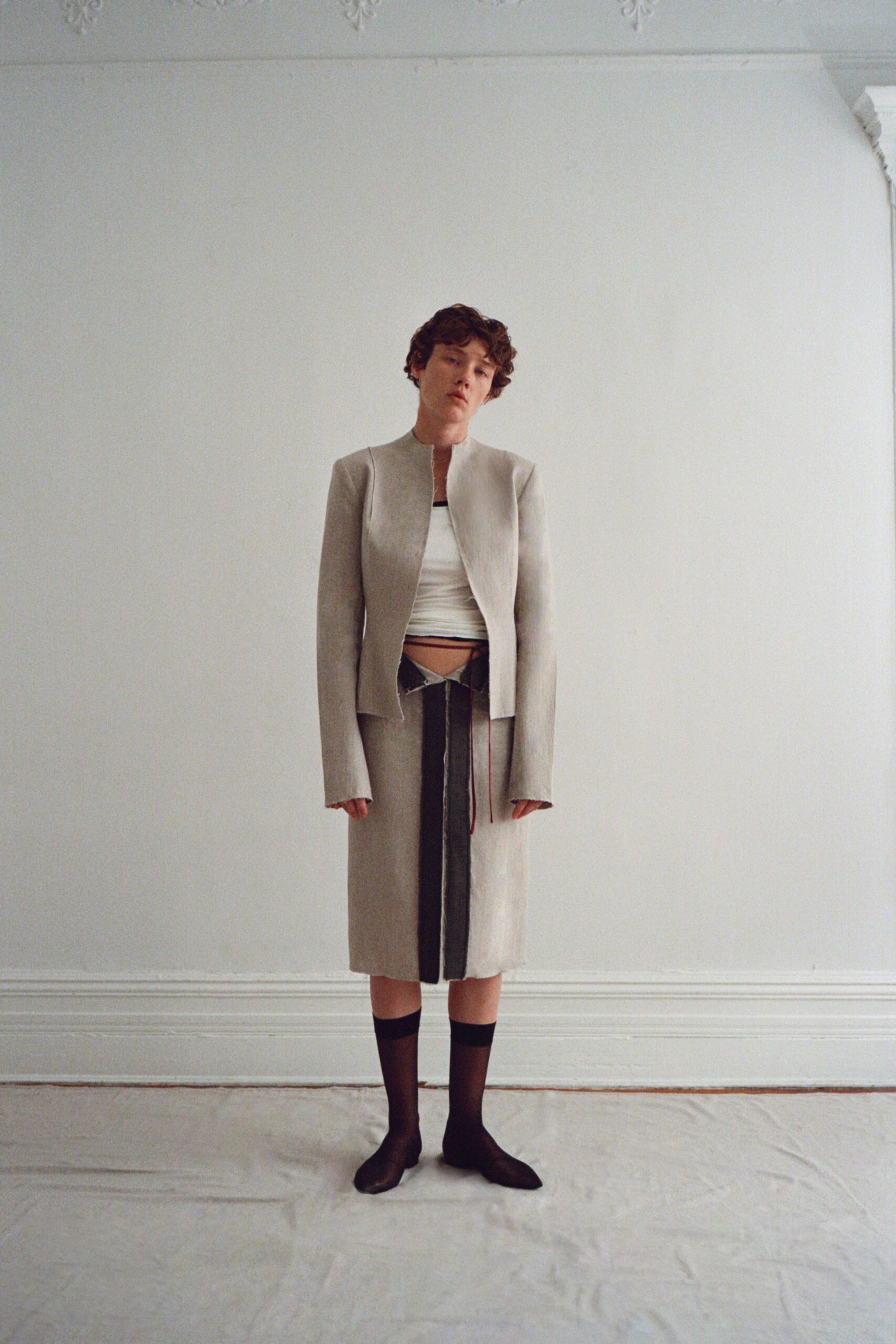 Pale grey collarless jacket, cut close with elongated sleeves past the wrist. White jersey tube top ruched tight across the torso. Matching midi skirt split by a dark center panel and dangling ties. Sheer black knee socks. In a drafty rented flat, still not unpacked after the move.