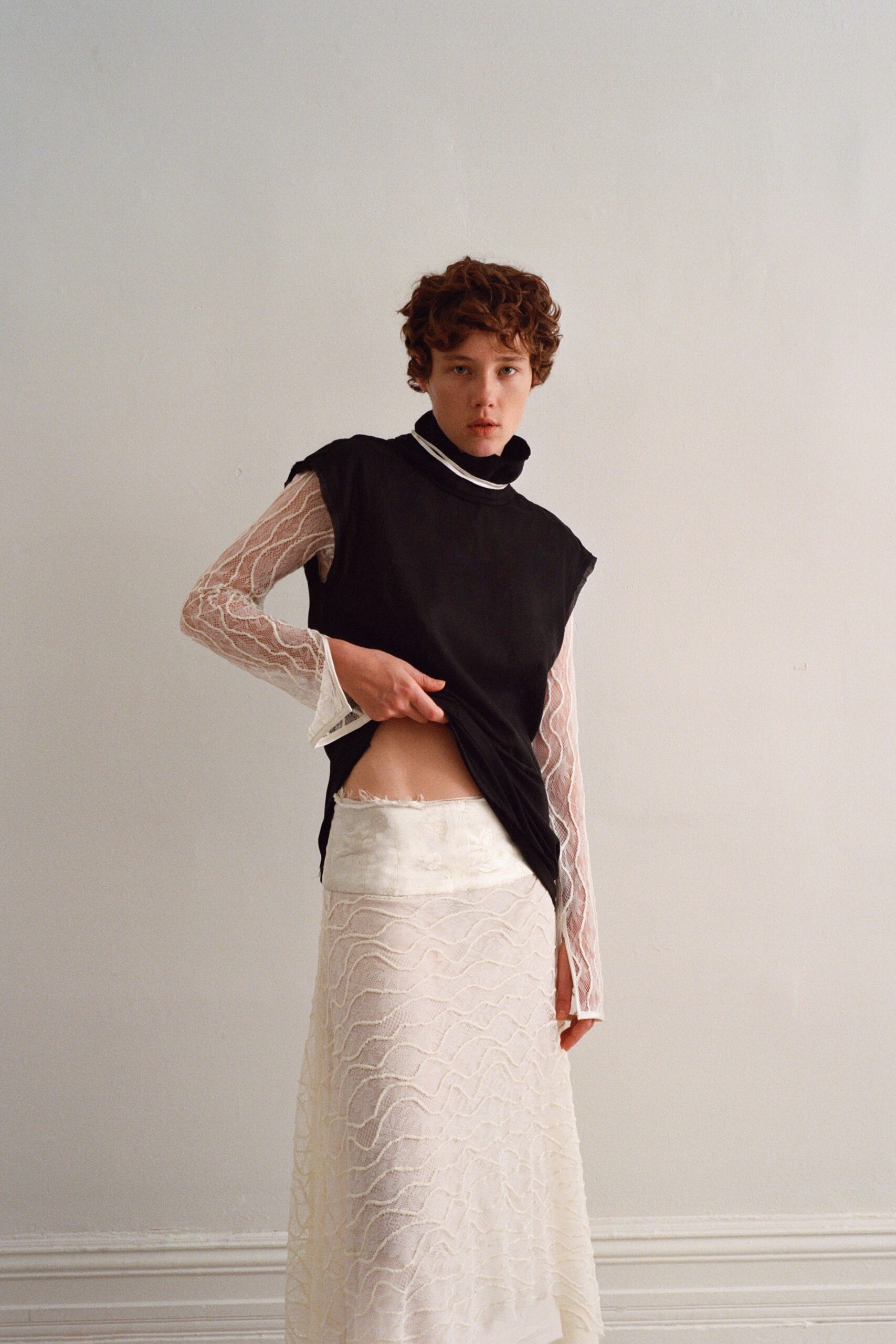 Black sleeveless jersey top, one side tugged up to show the waist. White sheer lace turtleneck with wavy raised cords and long sleeves. Ivory column skirt in matching corded lace, frayed waistband edge. Morning in a rented apartment, still not dressed for the meeting downstairs.